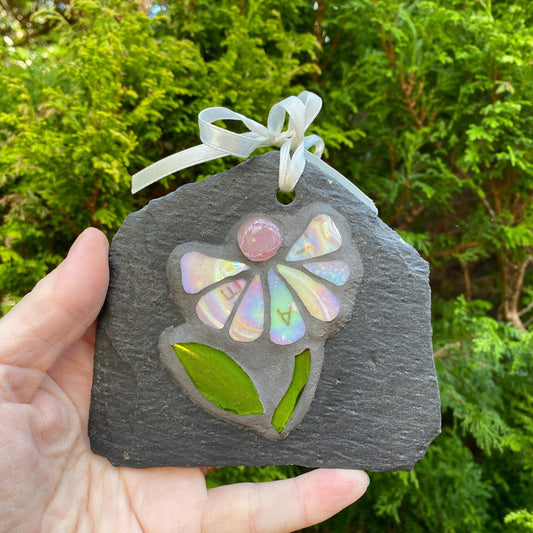 Small mosaic on slate handheld, showing a daisy with iridescent petals and a pink nugget and green leaves