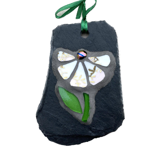 a small dark grey slate with a 5 petaled daisy with gilt on them, iridescent nugget and green glass stem and leave.  photographed on a white background
