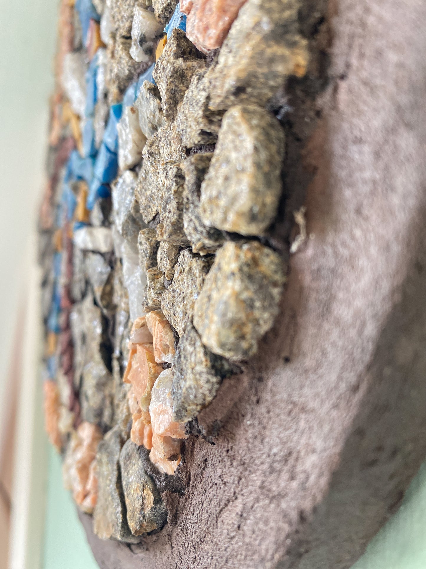 Detail of a circular mosaic made of various stones, with threads of blue and yellow plastic, and quartz representing stems, and pink toned stones for flower heads.  Brown painted edging. 