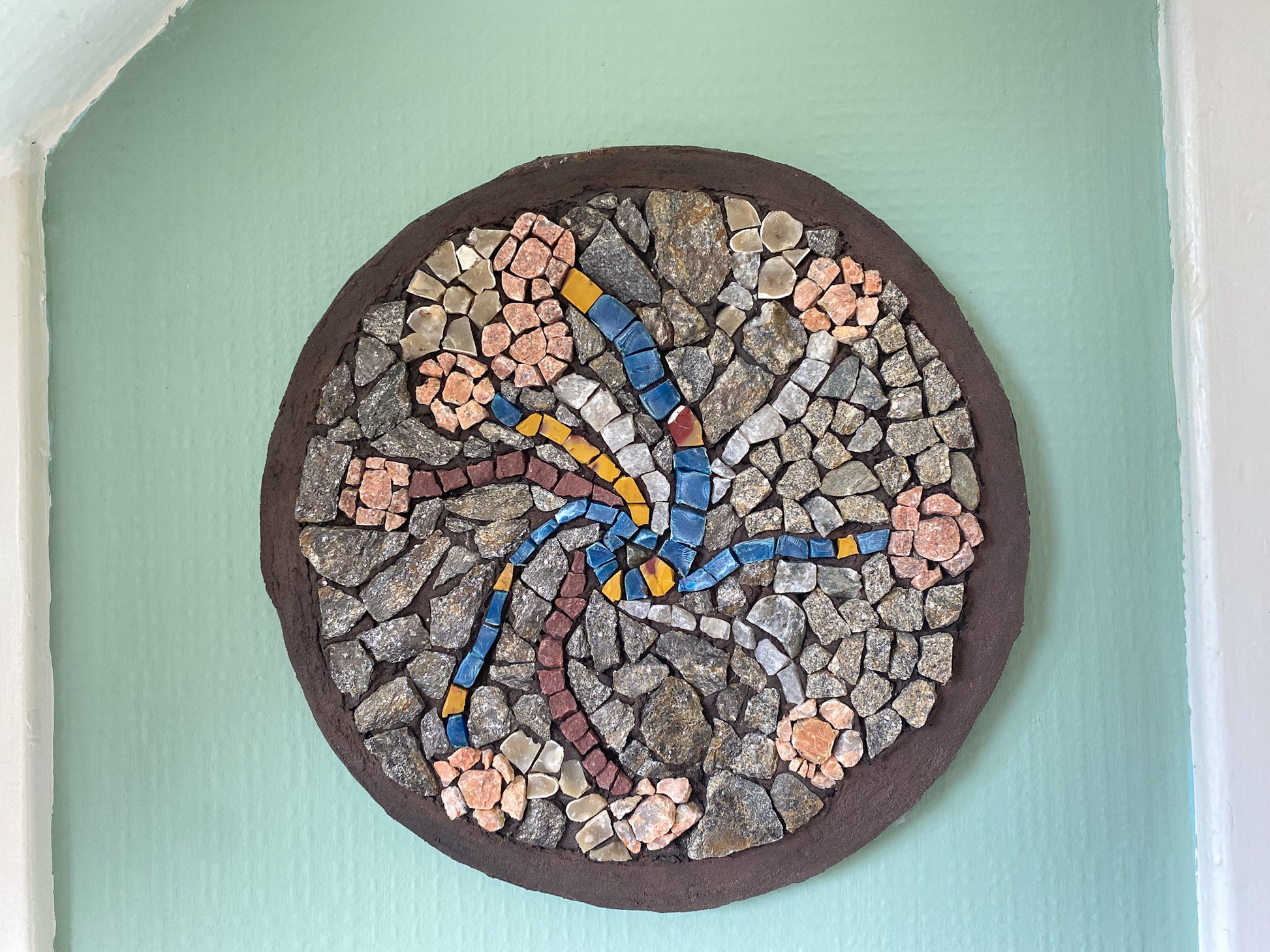 A circular mosaic made of various stones, with threads of blue and yellow plastic, and quartz representing stems, and pink toned stones for flower heads.  Brown painted edging.  Shown on a soft green wallpapered wall background