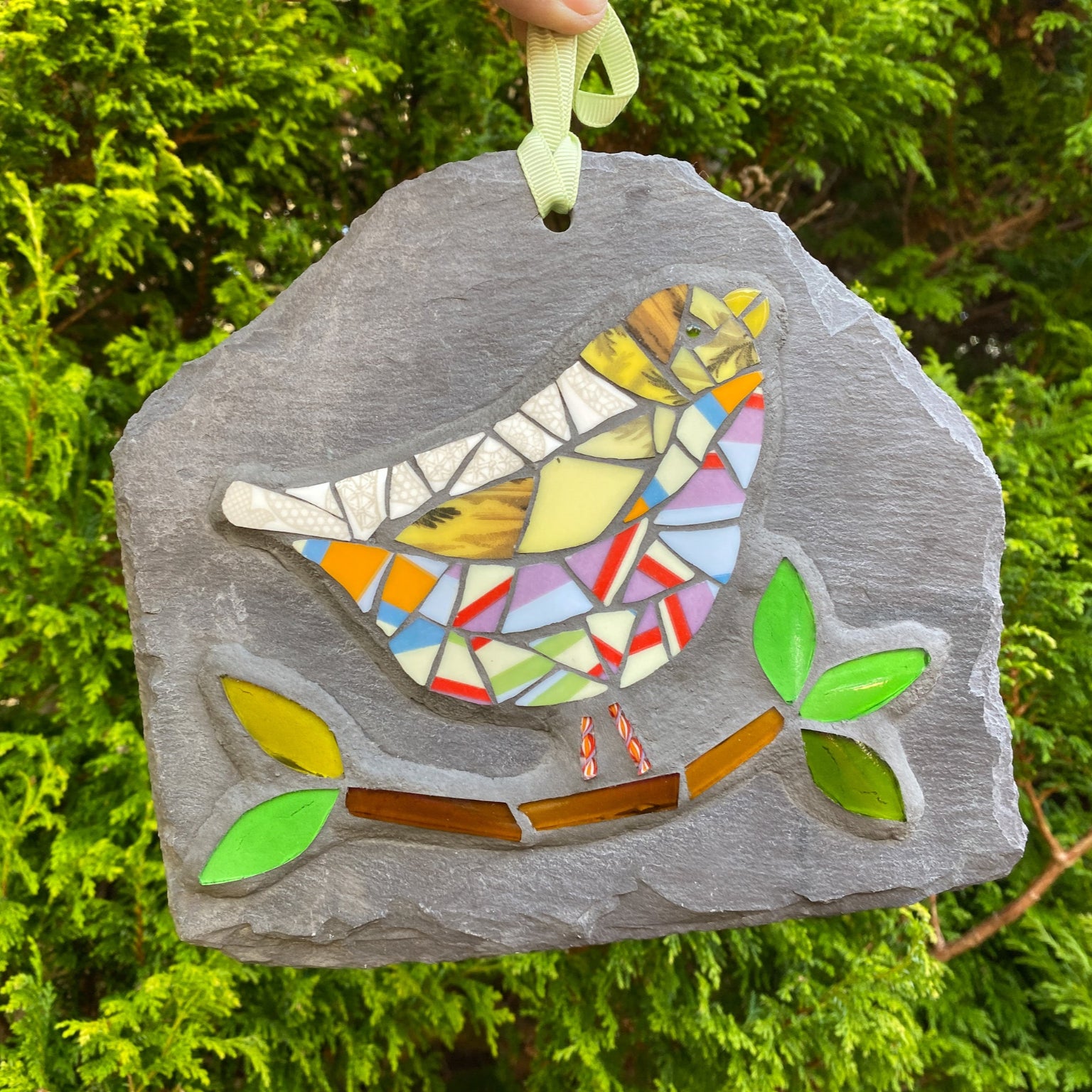 a colourful mosaic bird with lots of stripy tesserae with a branch and leaves in glass on a grey slate. photographed in front of an evergreen tree