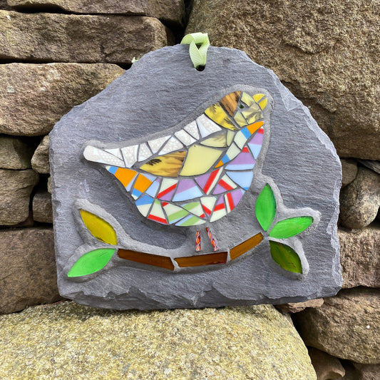 a colourful mosaic bird with lots of stripy tesserae with a branch and leaves in glass on a grey slate. photographed on a drystone wall