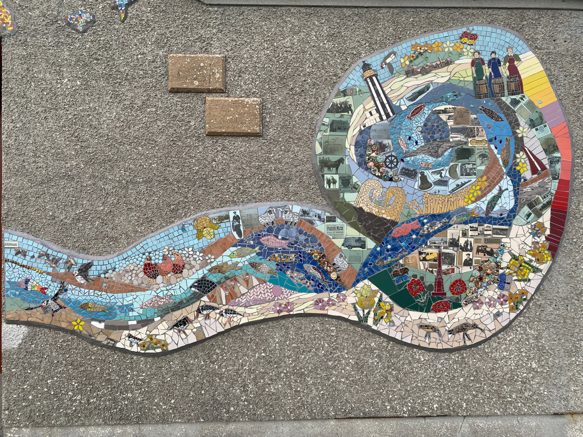 A large, detailed mosaic artwork depicting a winding river or stream with boats, figures, buildings, and natural elements, installed on a gray concrete surface.