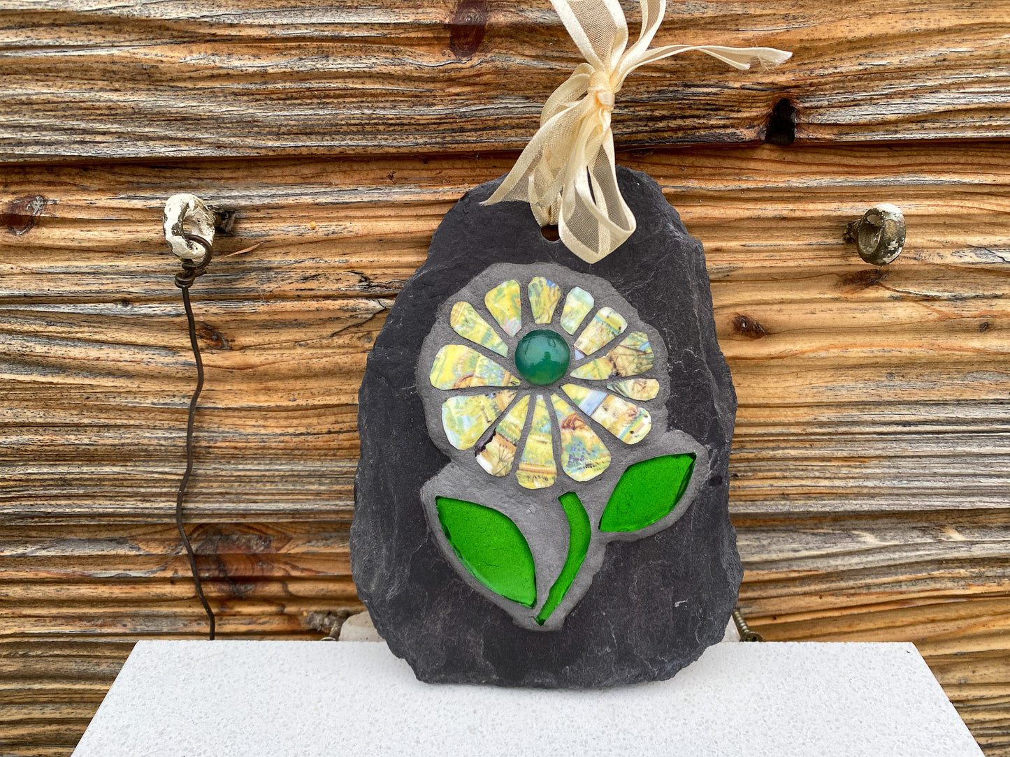 Mosaic flower with yellow patterned petals, and bright green leaves, on slate. Displayed against horizontals panelled wall