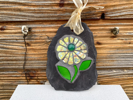 Mosaic flower with yellow patterned petals, and bright green leaves, on slate. Displayed against horizontals panelled wall