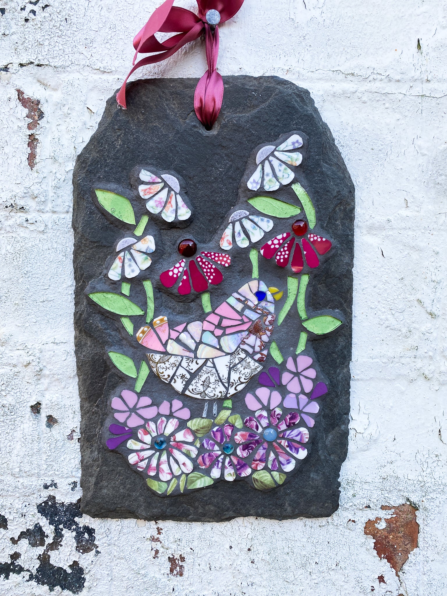 mosaic on slate of a pink gold and white bird surrounded by mosaic flowers in pink, hot pink and chintz tones.  leaves in pale green glass. shown on a distressed white brick wall