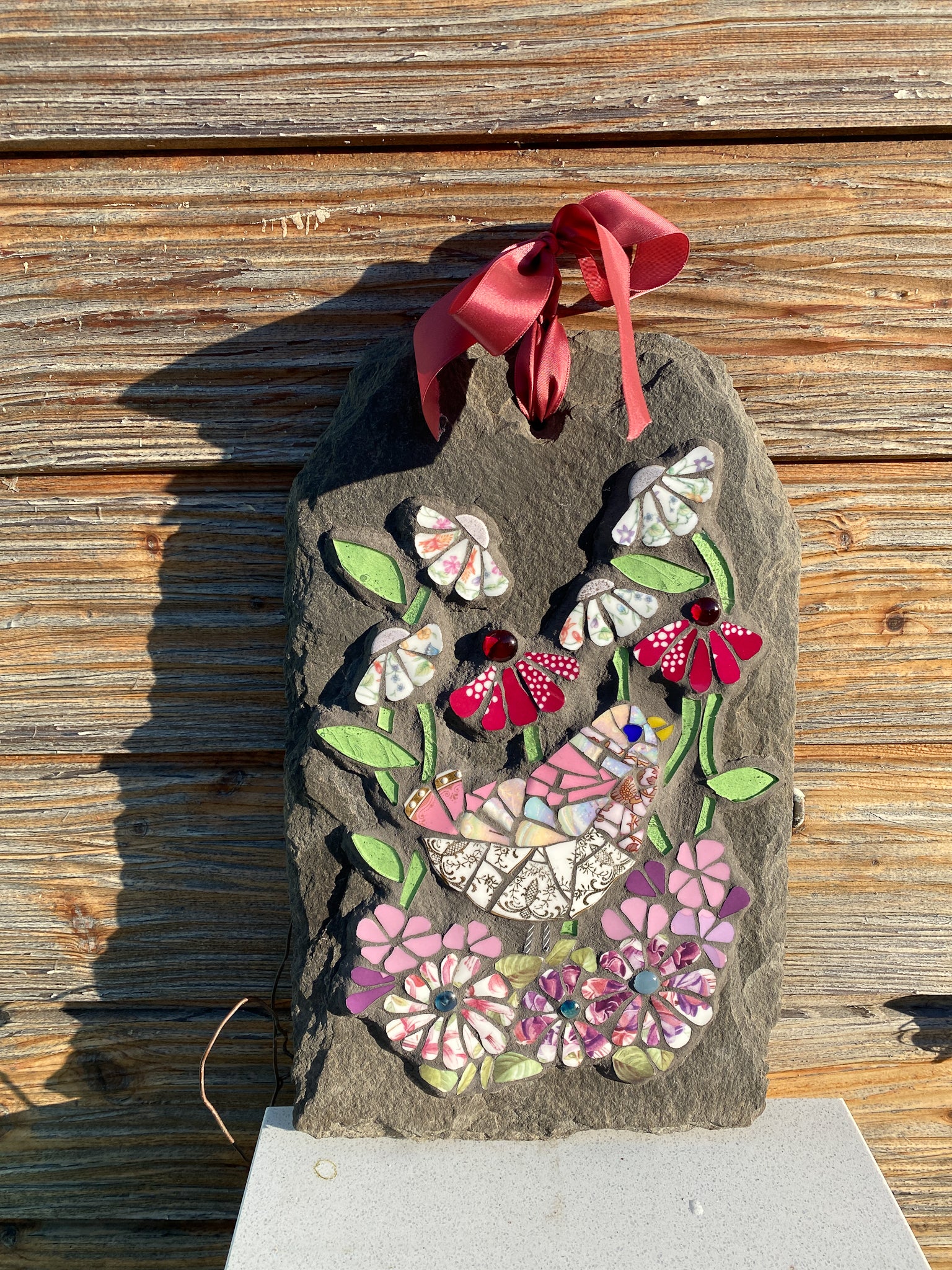 mosaic on slate of a pink gold and white bird surrounded by mosaic flowers in pink, hot pink and chintz tones.  leaves in pale green glass. Shown against a wooden background
