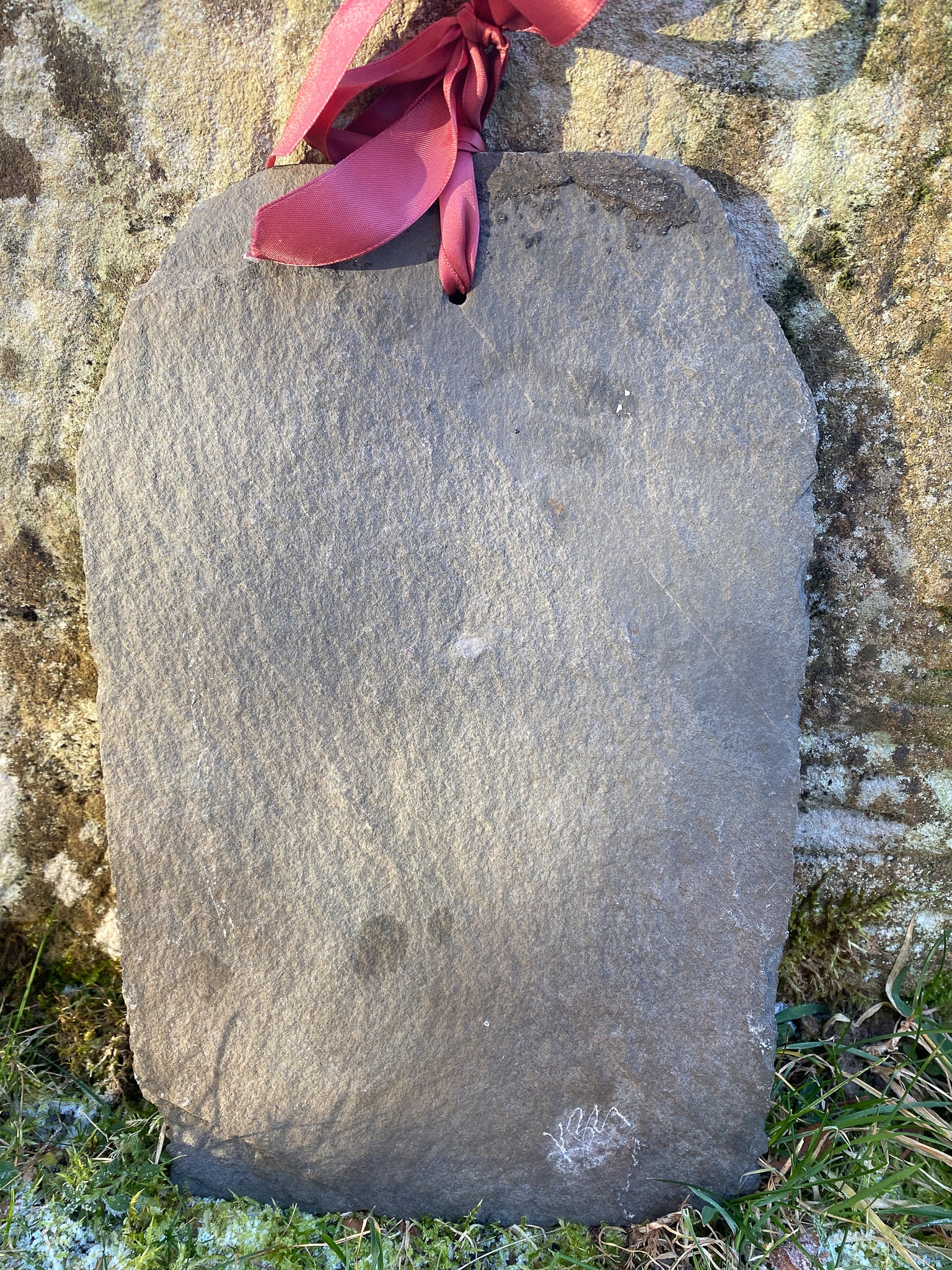 reverse of a cut roof slate showing a hole with maroon ribbon through, and artist signature