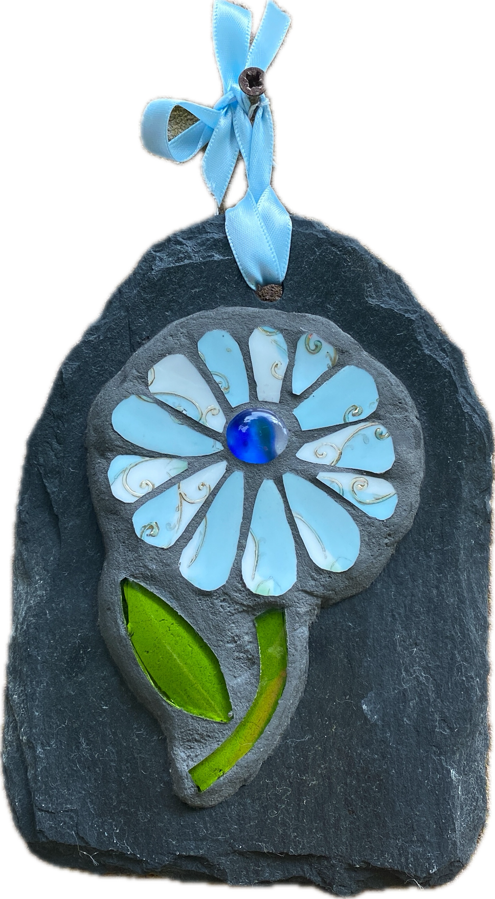 A handmade mosaic flower ornament mounted on a dark slate backing, featuring light blue glass petals, a blue glass center, green leaves, and a light blue ribbon loop at the top.