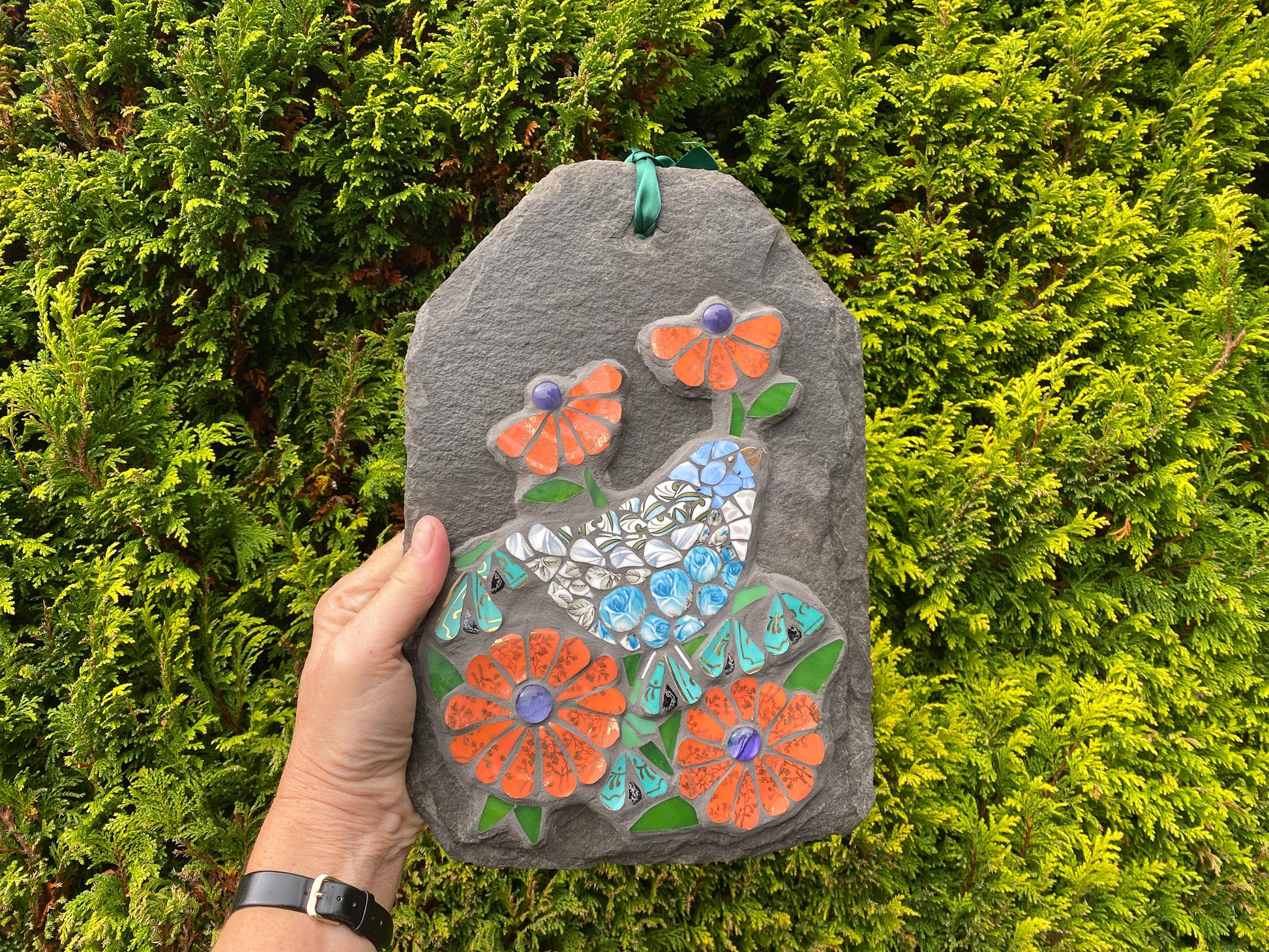 Decorative slate mosaic with a bird and flowers held in front of a green bush
