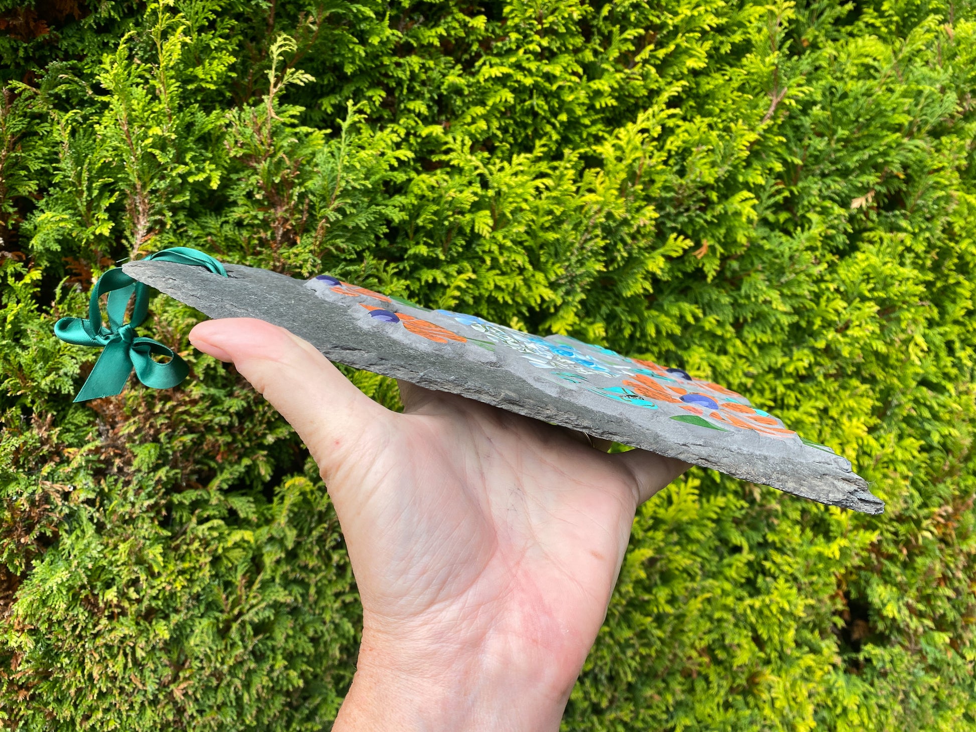 Hand holding a decorative slate with ribbons against a green bush background