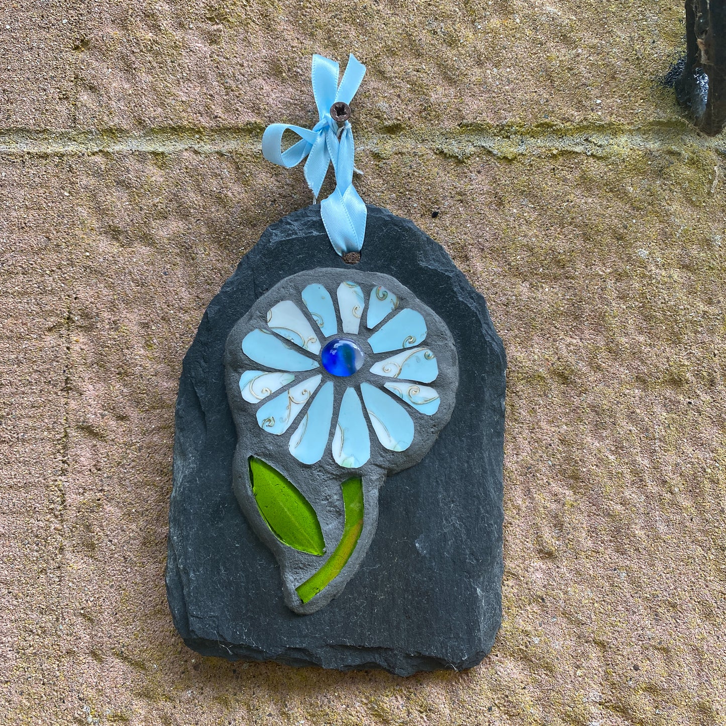 A handmade mosaic flower ornament hanging on a textured stone wall. The ornament is crafted from a dark slate base, featuring light blue glass petals, a central blue glass flower center, green glass leaves, and a light blue ribbon loop at the top.