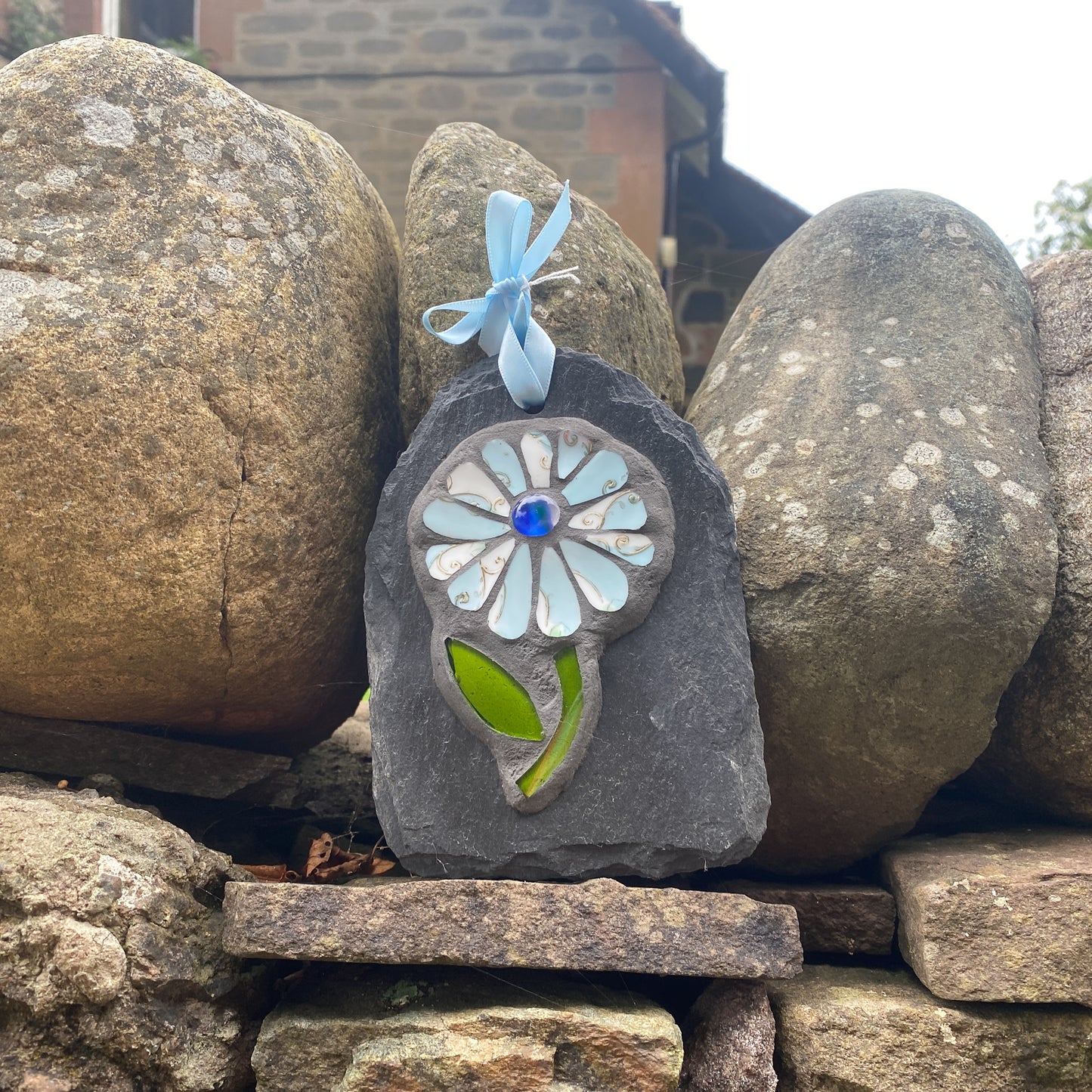 A decorative handmade stone plaque featuring a flower design made from light blue, white, and green glass pieces, with a blue glass center and a light blue ribbon loop at the top. The plaque is displayed outdoors on a rustic stone wall between large stones.