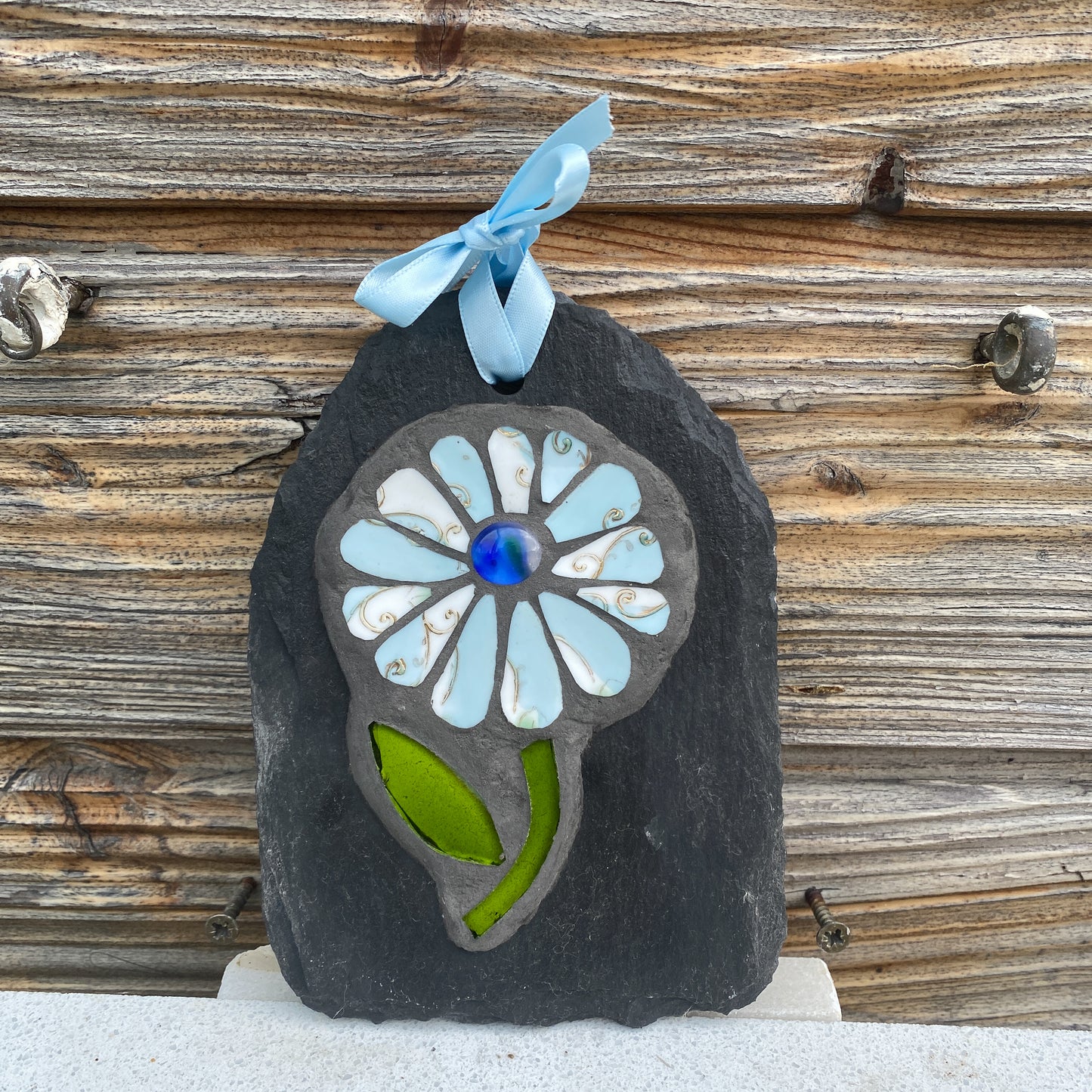 A handmade mosaic flower ornament on a dark slate plaque, featuring light blue glass petals, a blue glass center, and a green glass leaf, hanging from a light blue ribbon bow against a rustic wooden wall.