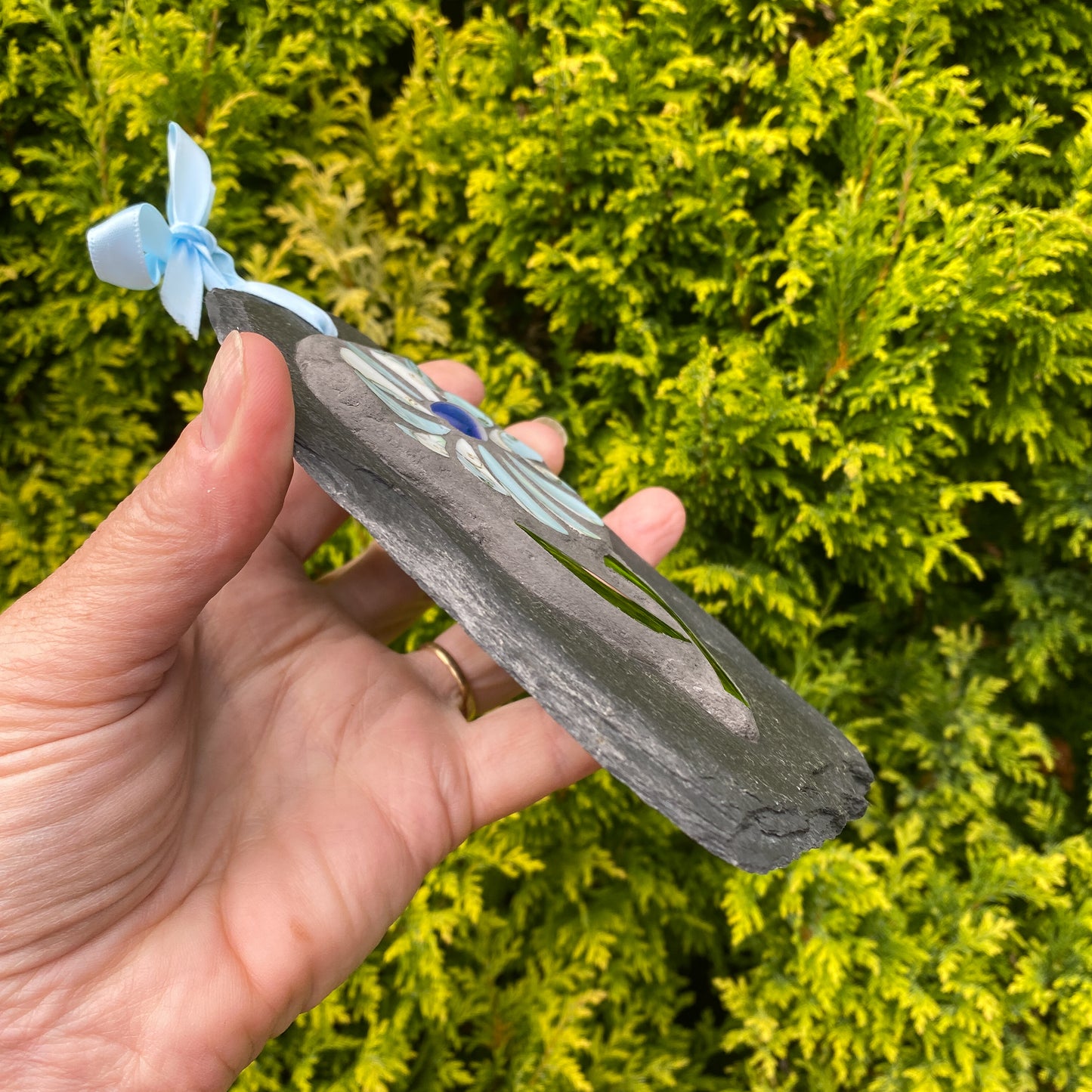 A hand holds a small decorative slate tag with a flower design and a light blue ribbon bow, set against a background of green foliage.