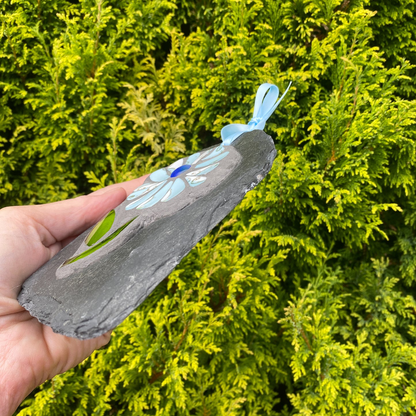 A hand holds a small slate garden ornament featuring a mosaic flower on a gray stone base, set against a background of bright green conifer branches.