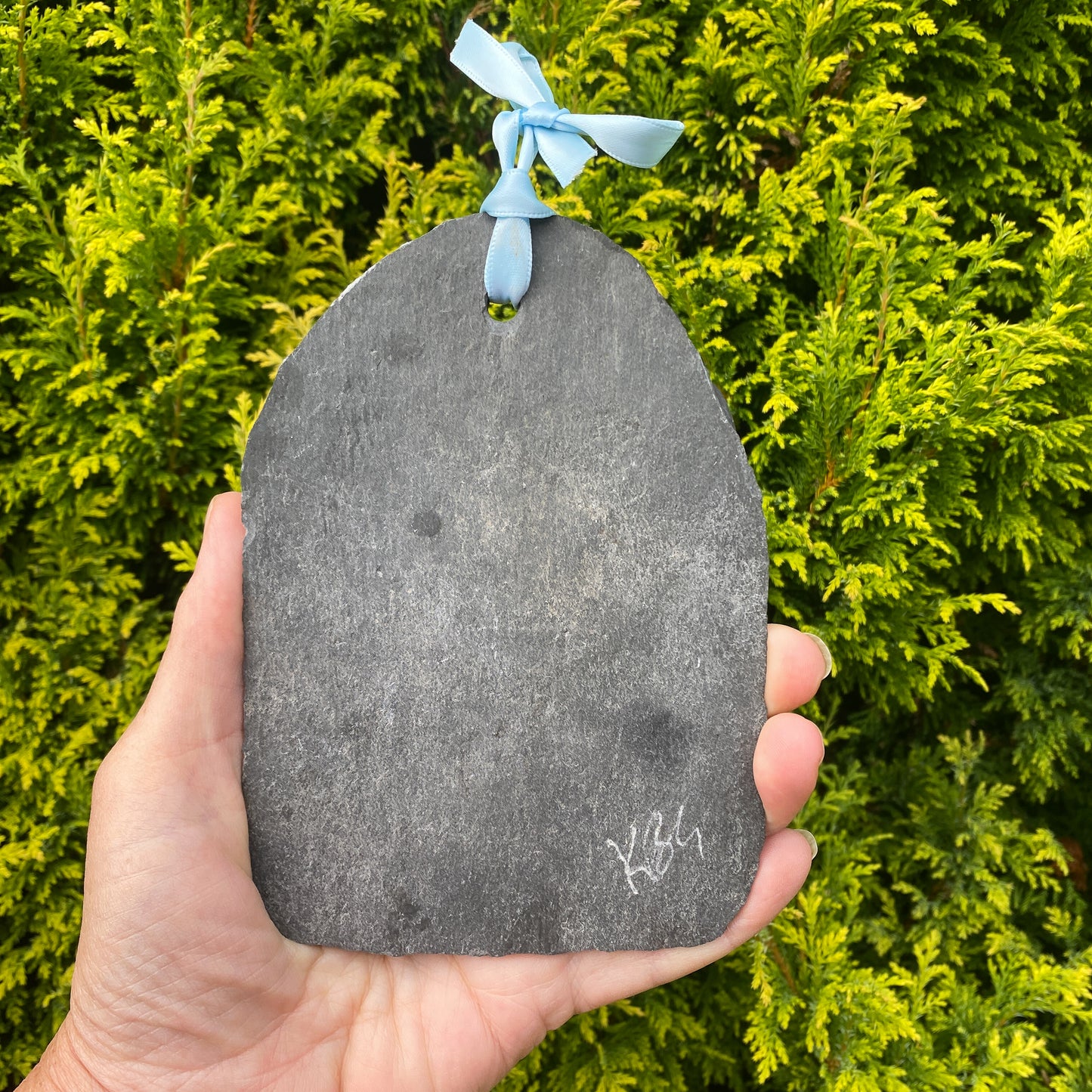 A hand holds a dark gray slate ornament shaped, featuring a light blue ribbon bow at the top and a white painted symbol near the bottom, set against a background of green foliage.