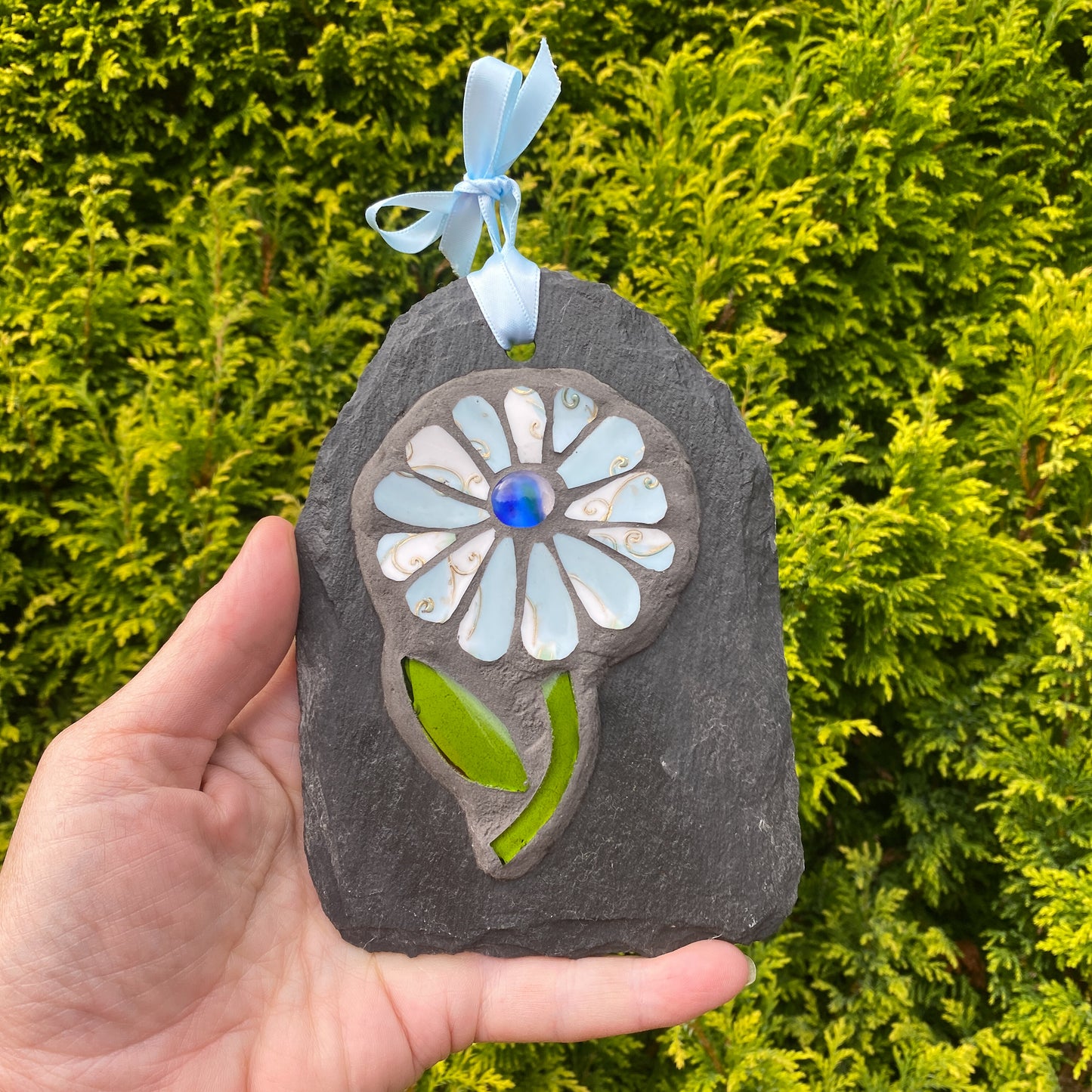 A hand holds a decorative garden ornament shaped like a flower, featuring a gray stone base, a white and light blue glass flower with a blue center, green glass leaves, and a light blue ribbon at the top, set against a background of green foliage.
