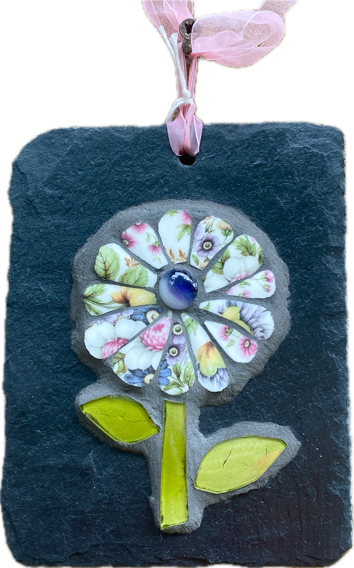 A square dark slate wall hanging featuring a raised flower design made from colorful patterned stone petals, a blue glass center, and two bright green leaves, suspended by a pink ribbon against a white background.