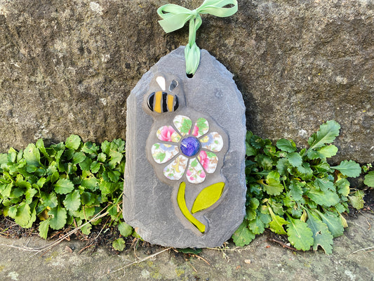 mosaic flower with chintz petals, purple glass nugget at its centre, and green stem.  there is a mosaic bee beside the flower.  on a grey roof slate, shown on a stone step with foliage around