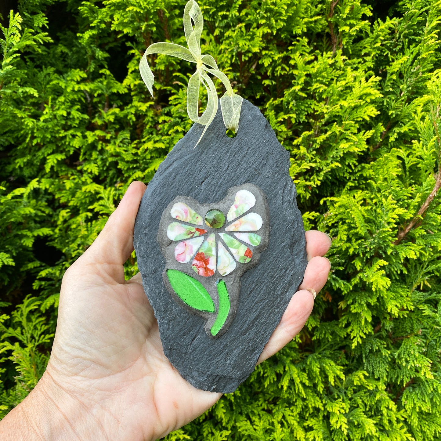 Hand holding a decorative slate with a floral design against a green bush background
