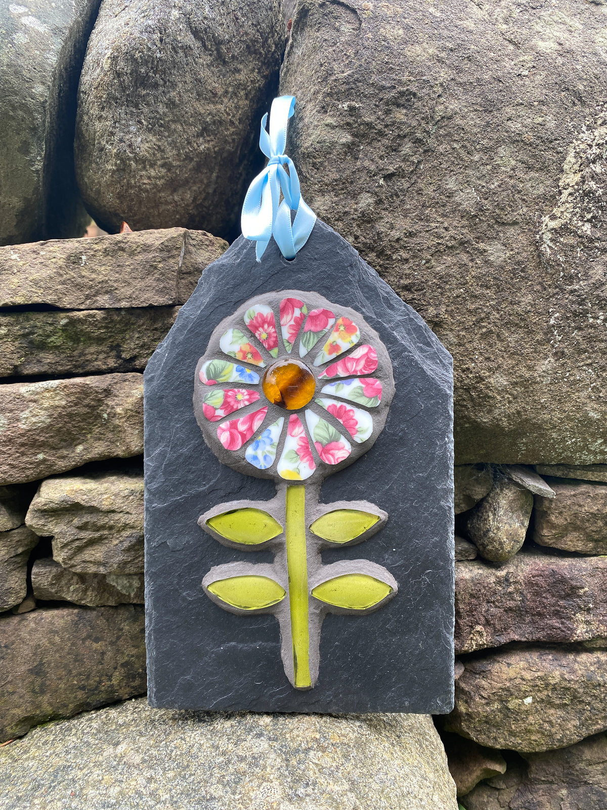 A handmade mosaic flower ornament on a dark slate plaque, featuring a colorful floral design with a light blue ribbon loop at the top, set against a rustic stone wall background.