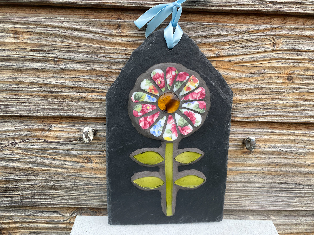 A handmade decorative wall hanging made from a dark slate plaque featuring a colorful floral design with pink, blue, and yellow mosaic petals, a central amber glass circle, and lime green leaves. It hangs from a light blue ribbon against a rustic wooden wall.