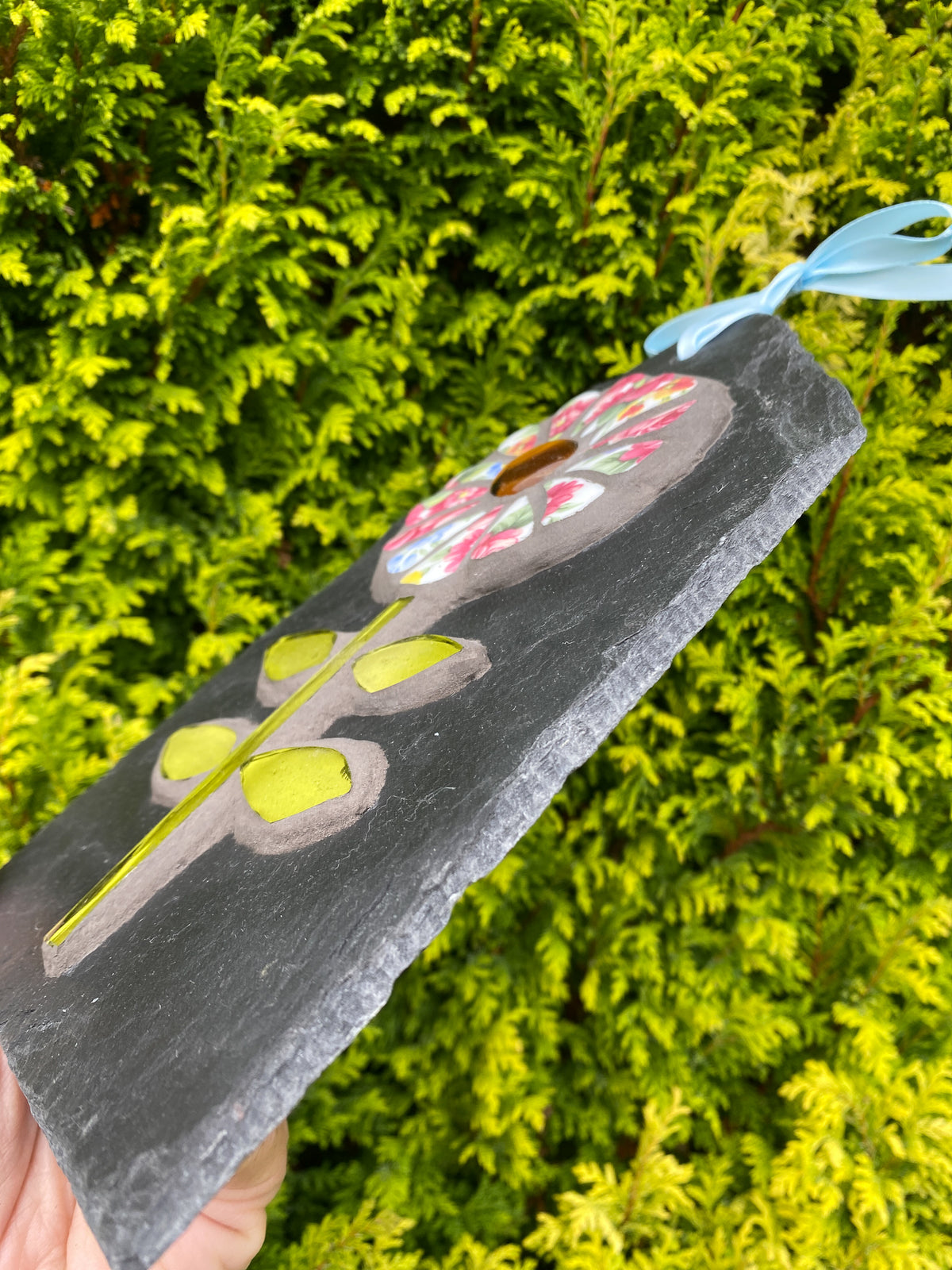 A hand holds a rectangular dark slate plaque featuring a mosaic flower design with green leaves and a colorful circular motif, set against a background of bright green foliage.