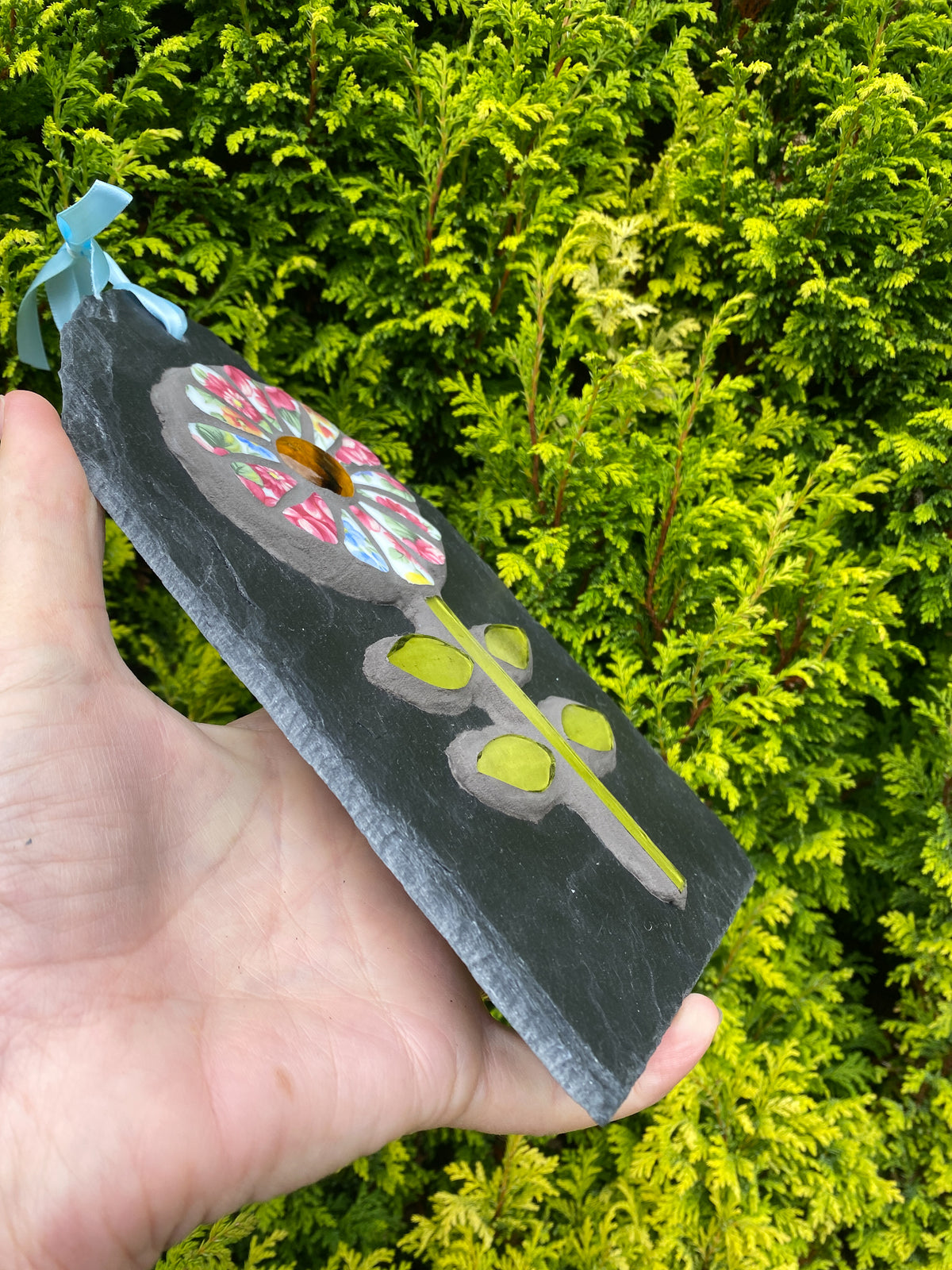 A hand holds a rectangular black slate ornament featuring a mosaic flower design with a light blue ribbon loop, set against a background of bright green foliage.