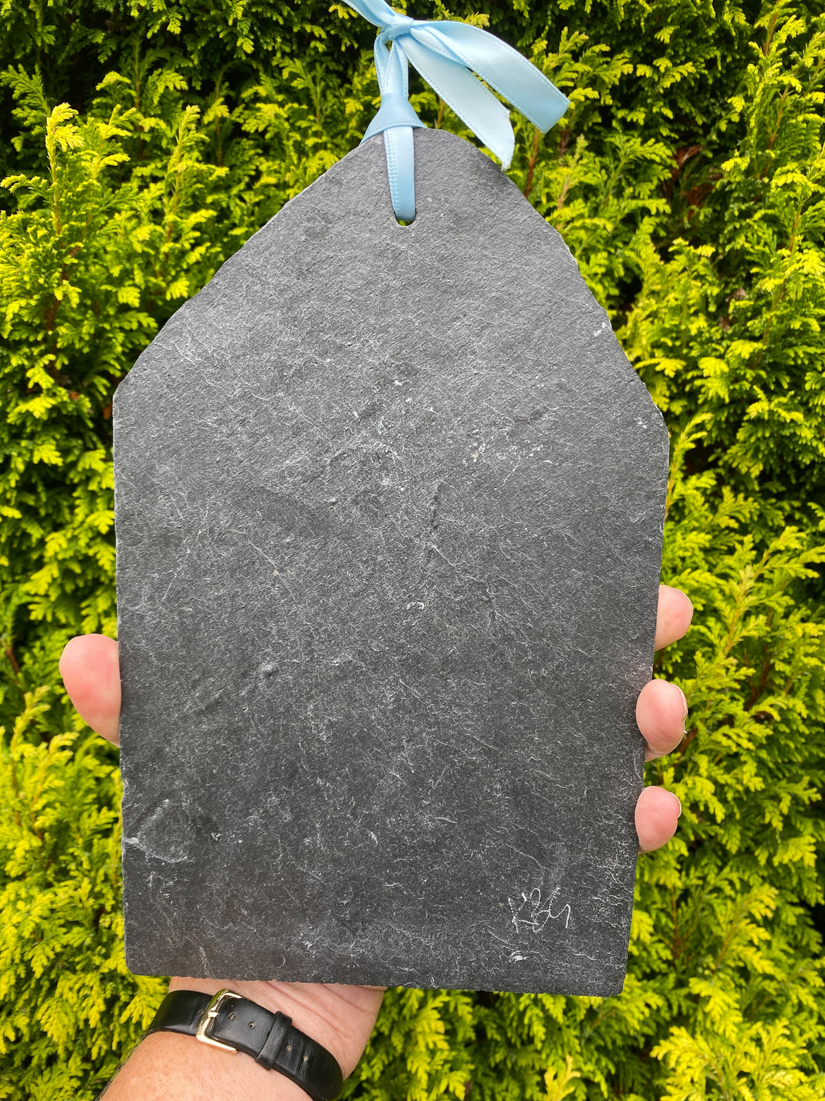 A hand holds a dark gray slate ornament shaped like a house, tied at the top with a light blue ribbon, against a background of green foliage.