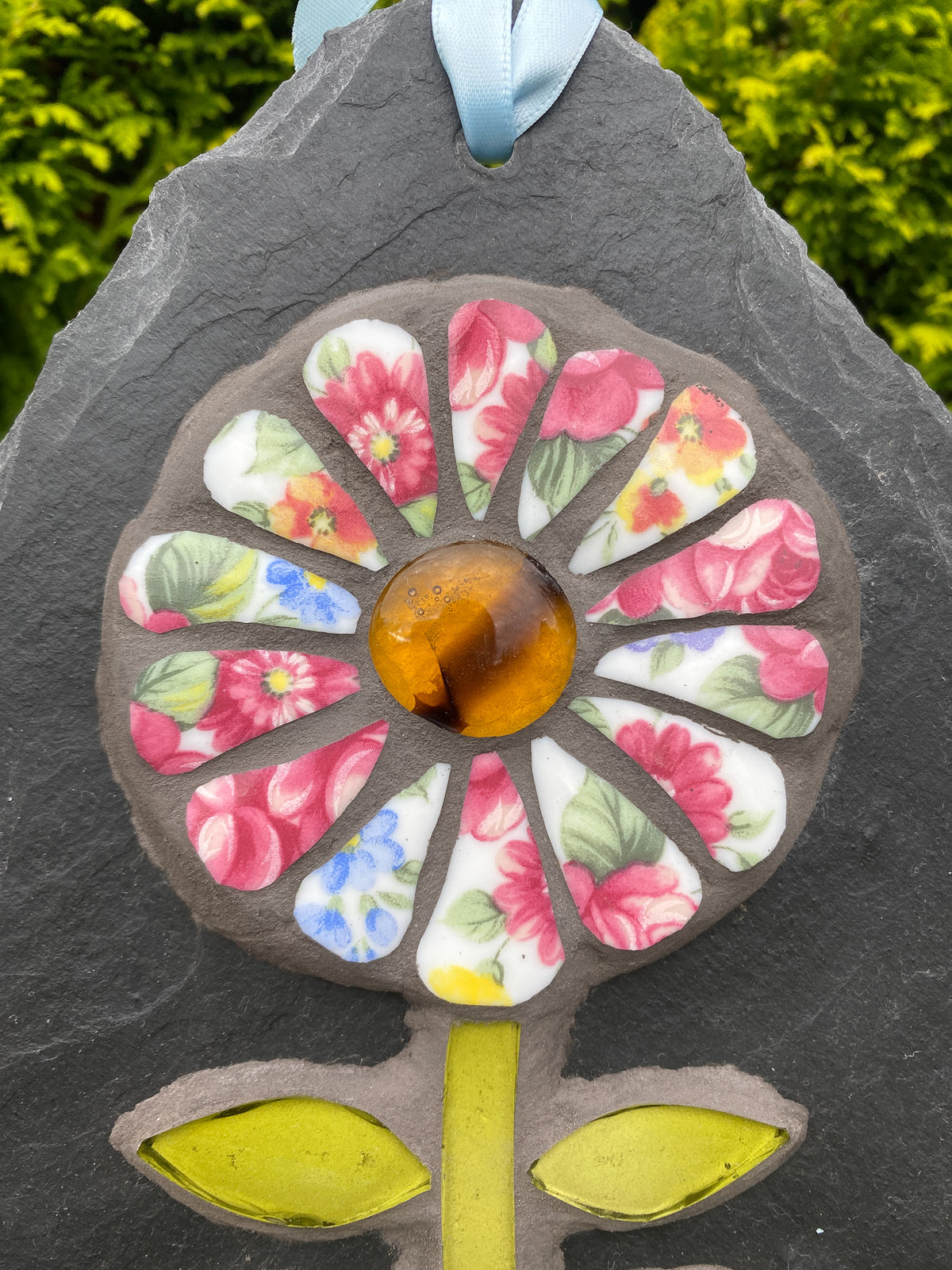 A close-up of a decorative flower-shaped garden ornament or plaque mounted on a dark slate backing. The flower has a glossy amber-yellow center and multicolored floral petal panels in pink, blue, yellow, and white, with a light blue hanging ribbon at the top.