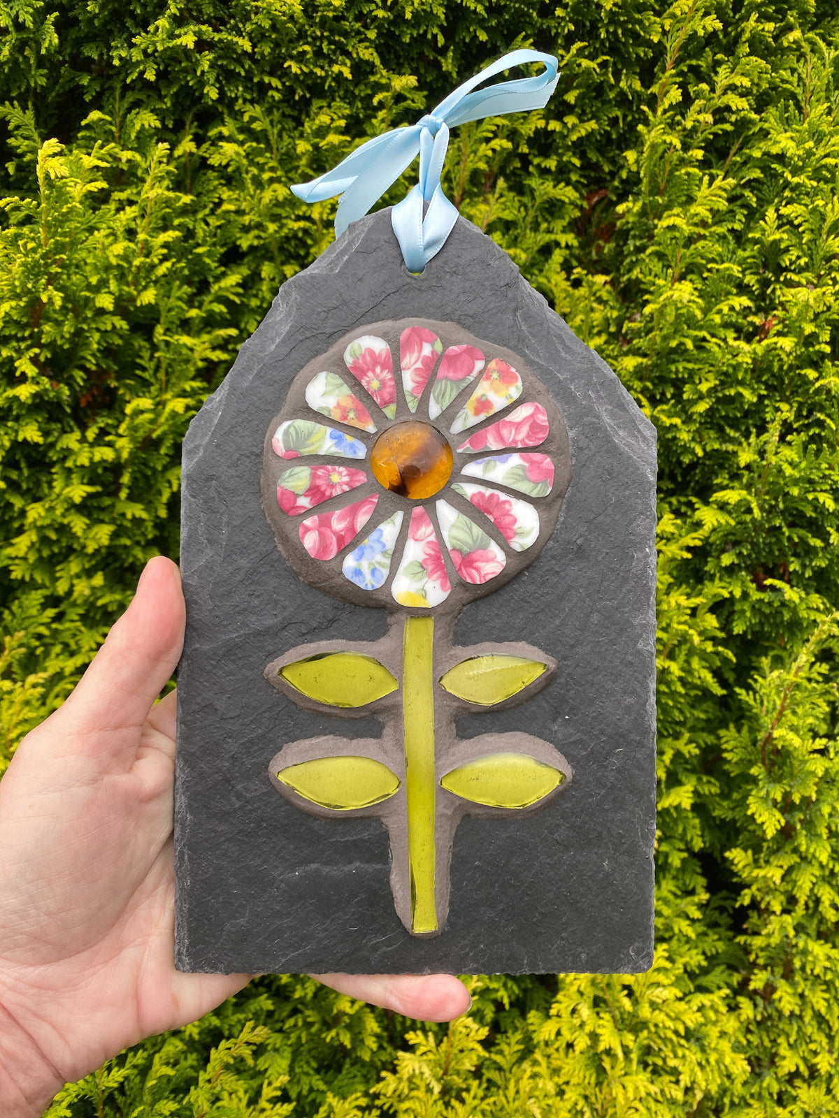 A hand holds a decorative slate plaque shaped like a house with a colorful mosaic flower on it and a light blue ribbon hanging from the top. The background is a dense green hedge.