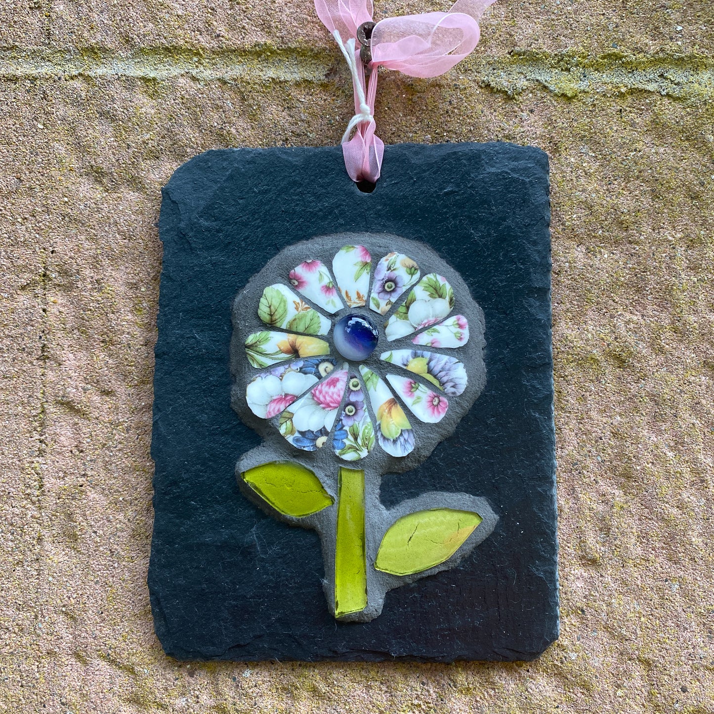 A square black slate wall hanging ornament featuring a colorful mosaic flower with a pink ribbon loop at the top, displayed against a textured beige stone wall.