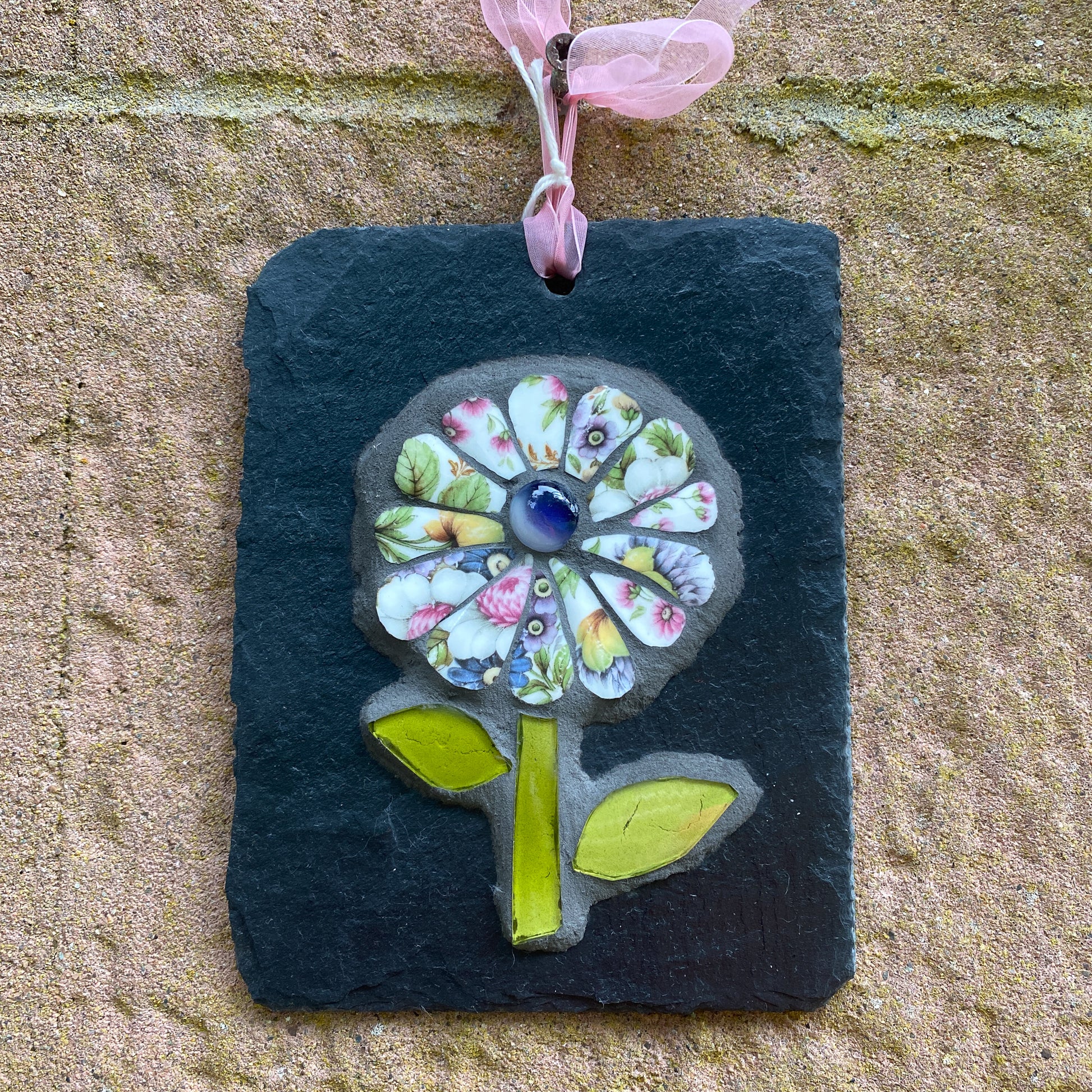 A square black slate wall hanging ornament featuring a colorful mosaic flower with a pink ribbon loop at the top, displayed against a textured beige stone wall.