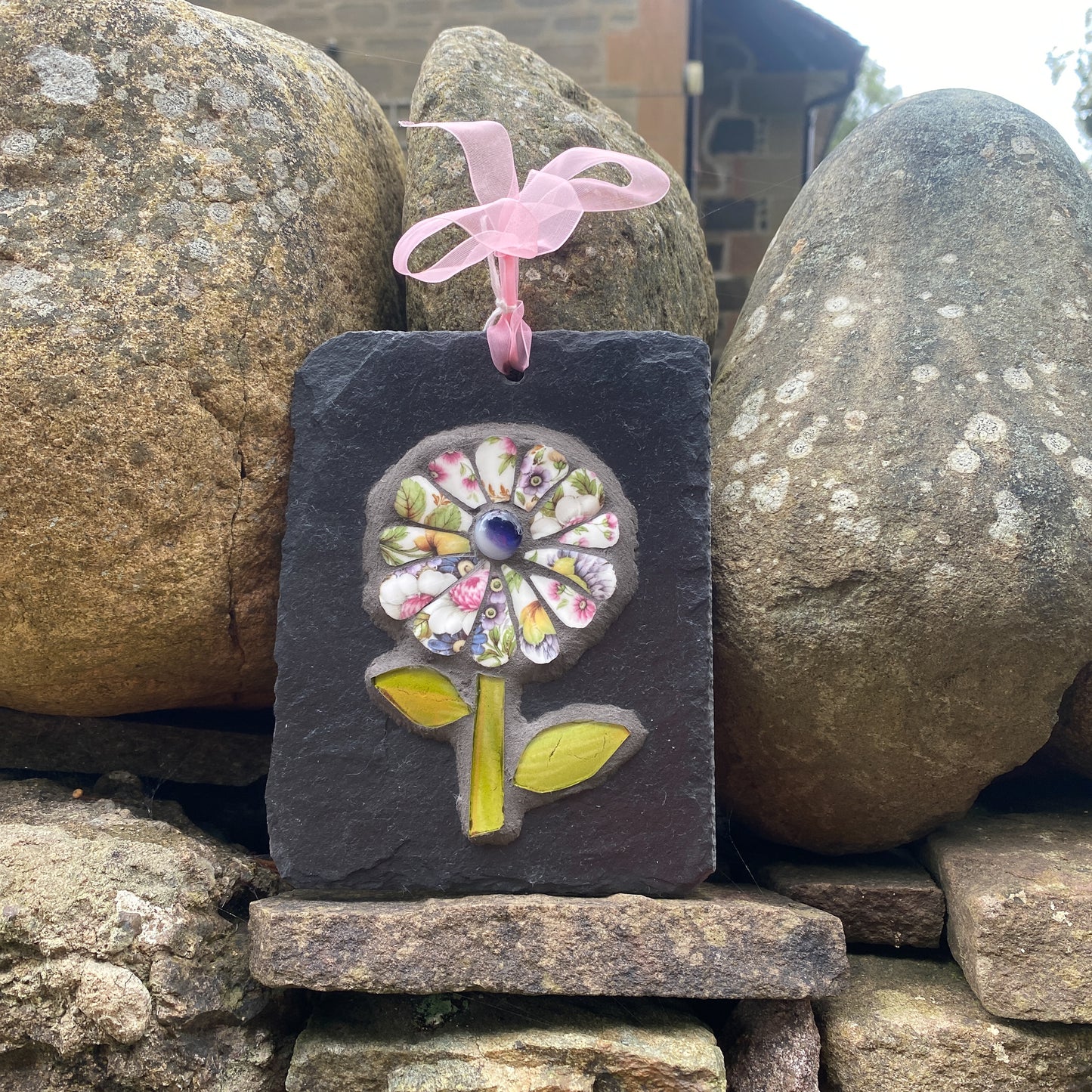 A rectangular dark slate wall hanging ornament featuring a colorful mosaic flower design with a pink ribbon loop at the top, displayed outdoors between large natural rocks.