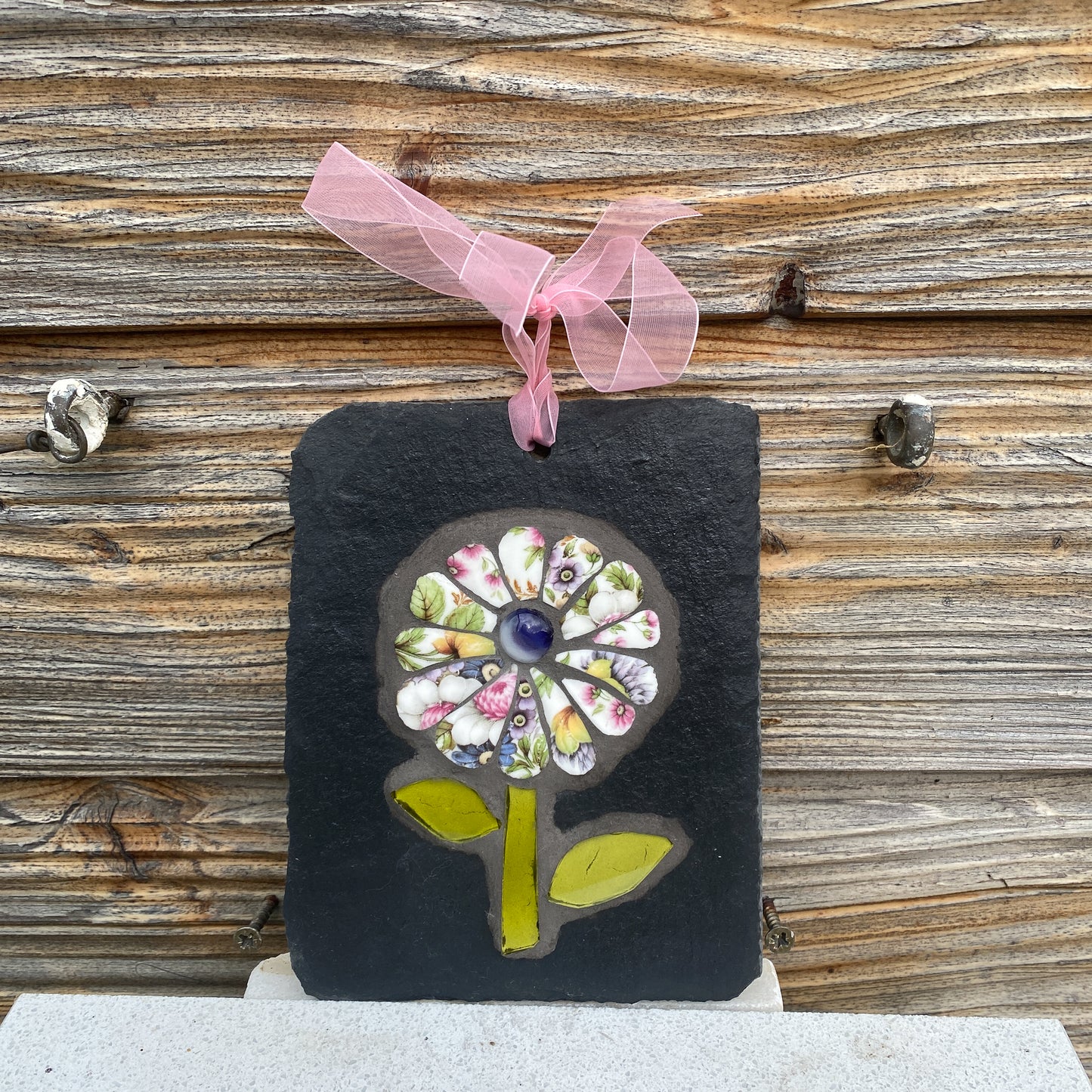 A handmade rectangular black slate wall hanging ornament featuring a colorful mosaic flower design with a pink sheer ribbon loop at the top, displayed against a rustic wooden plank background.