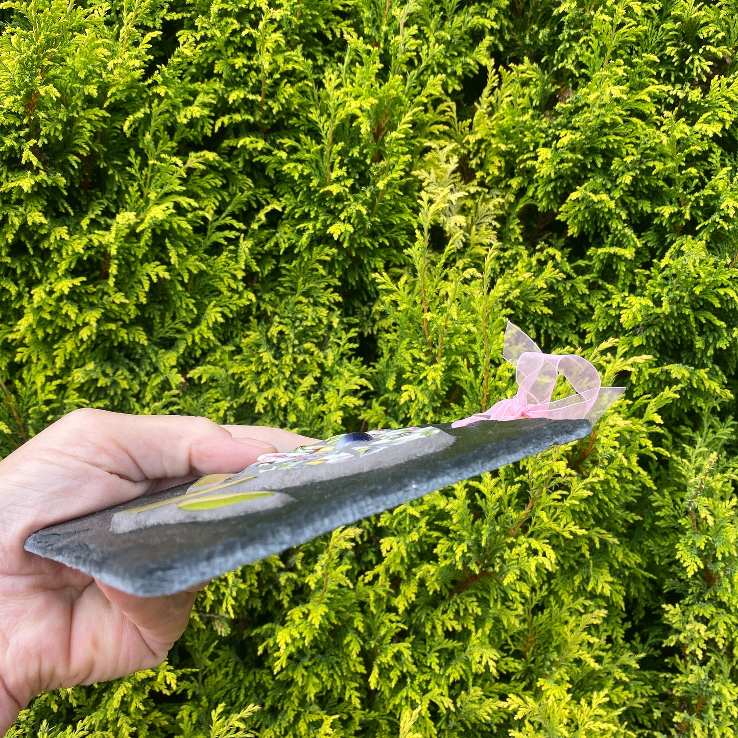 A hand holding a rectangular dark gray slate tile outdoors in front of a dense green hedge. The tile has colorful mosaic details and a small pink translucent ribbon tied to one corner.