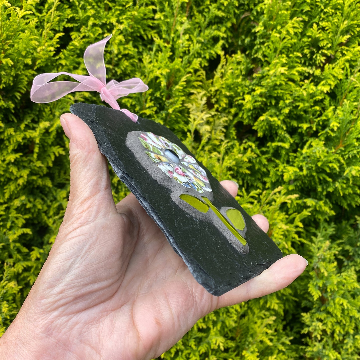 A hand holds a rectangular black slate plaque featuring a colorful mosaic floral design and a green glass accent, tied with a pink ribbon, against a background of green foliage.