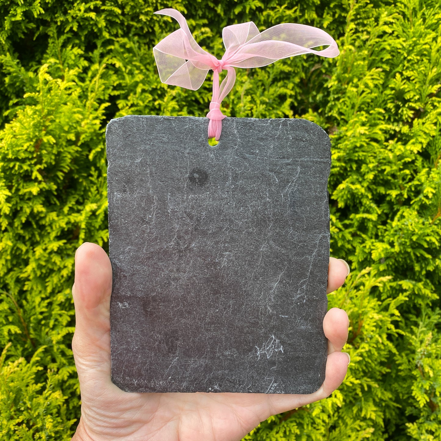 A rectangular dark gray slate hanging ornament with rounded corners and a pink sheer ribbon loop at the top, held in a hand against a background of green foliage.