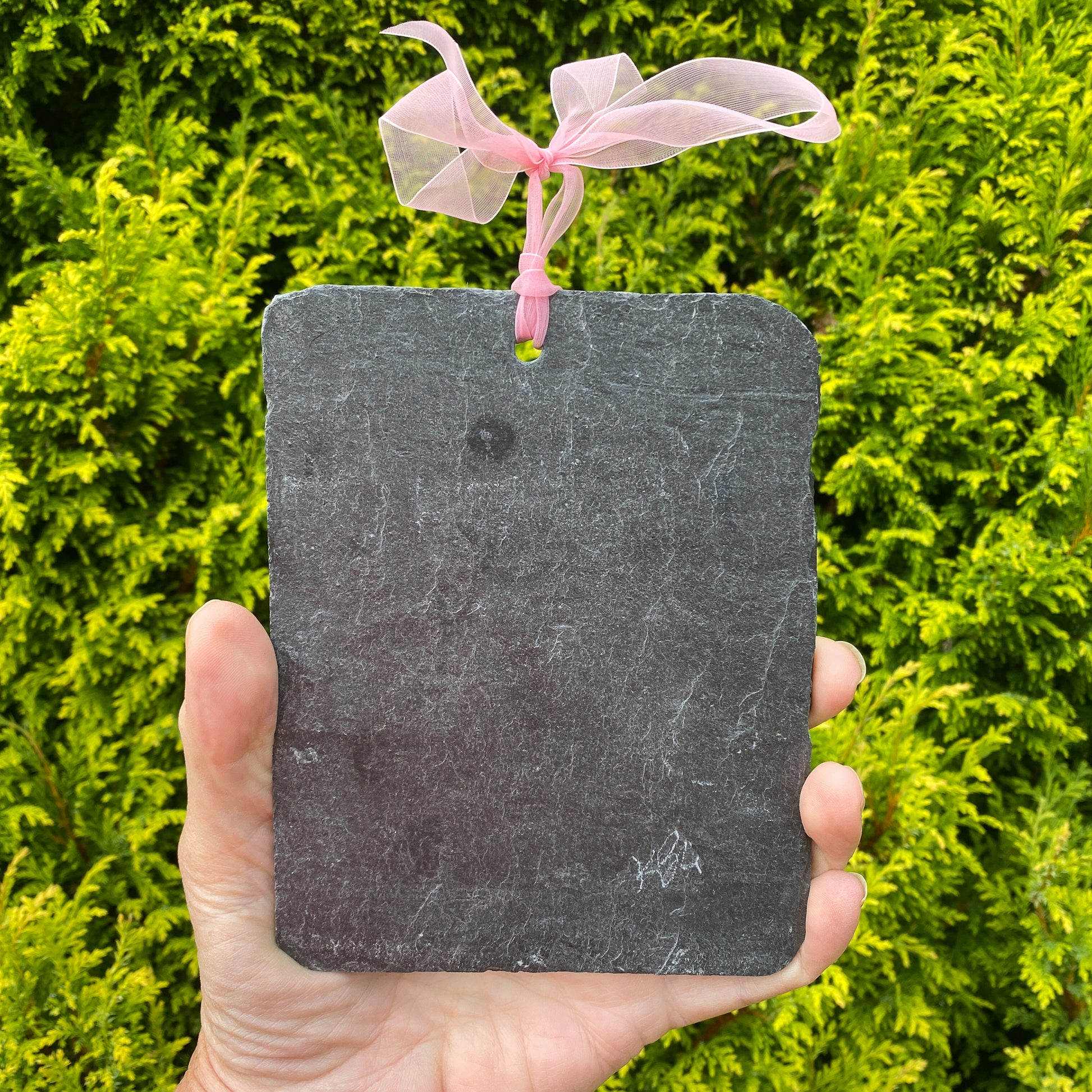A rectangular dark gray slate hanging ornament with rounded corners and a pink sheer ribbon loop at the top, held in a hand against a background of green foliage.