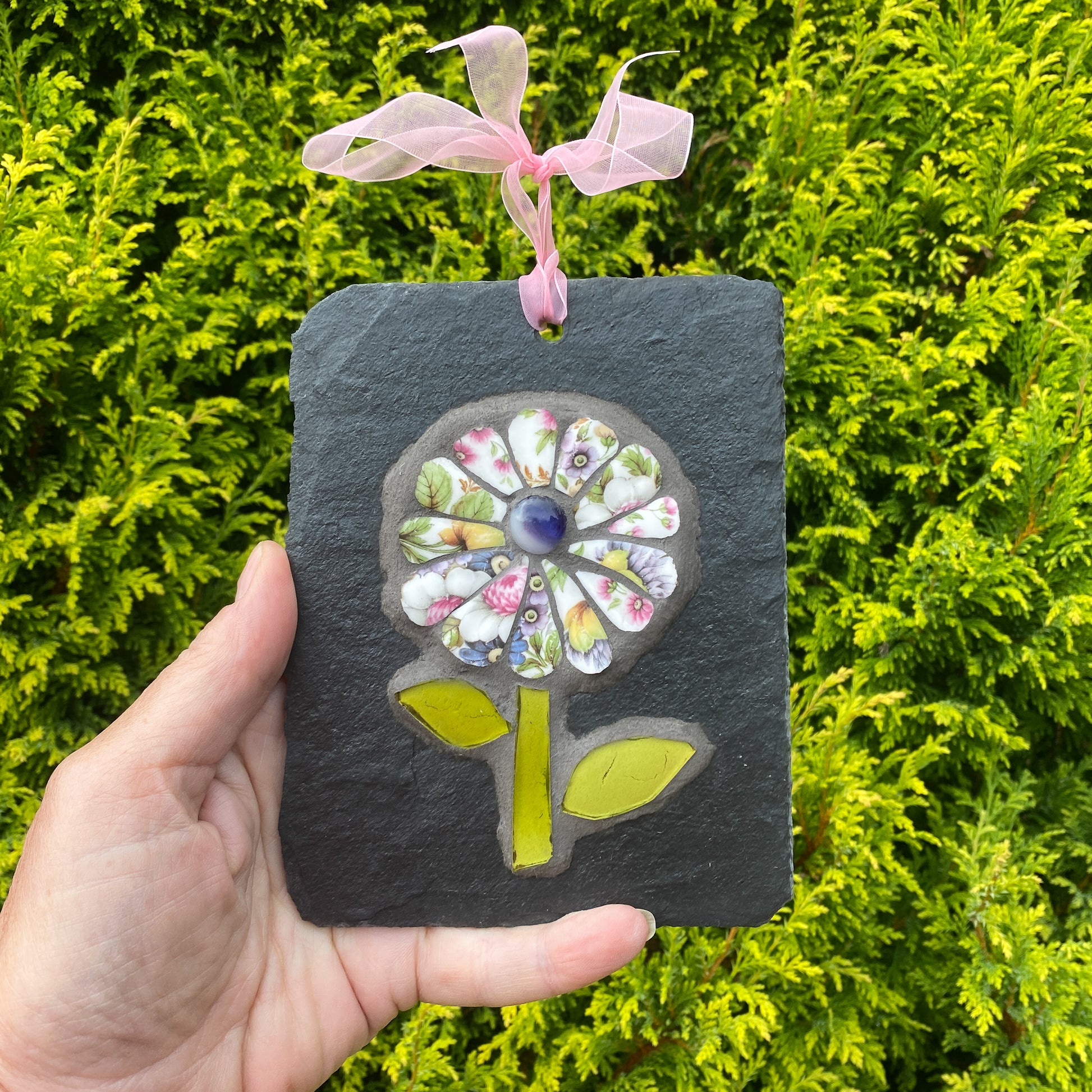 A hand holds a square dark slate plaque featuring a colorful mosaic flower design with a pink ribbon loop at the top, set against a background of green foliage.