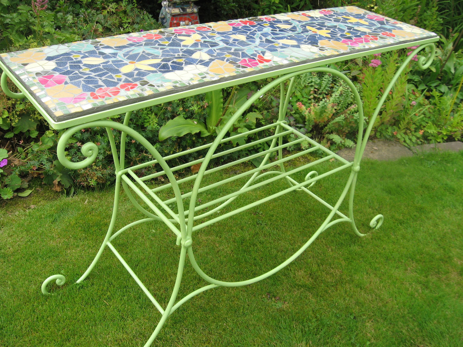 mosaic displayed in a garden.  The table has lime green paint on the metal, and the top has a mosaic of flowers with blue background