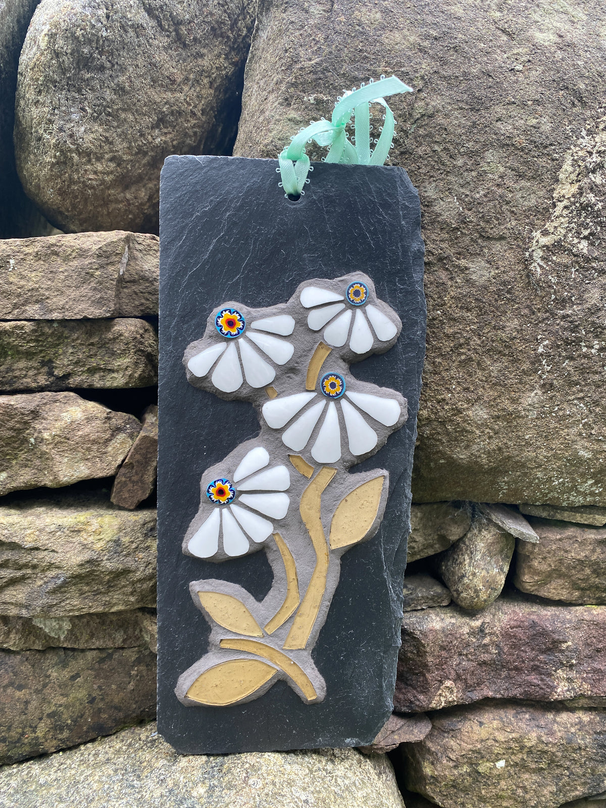 A rectangular dark slate wall plaque featuring raised white daisy flowers mosaic with yellow and blue centers, set against a rustic stone wall background.