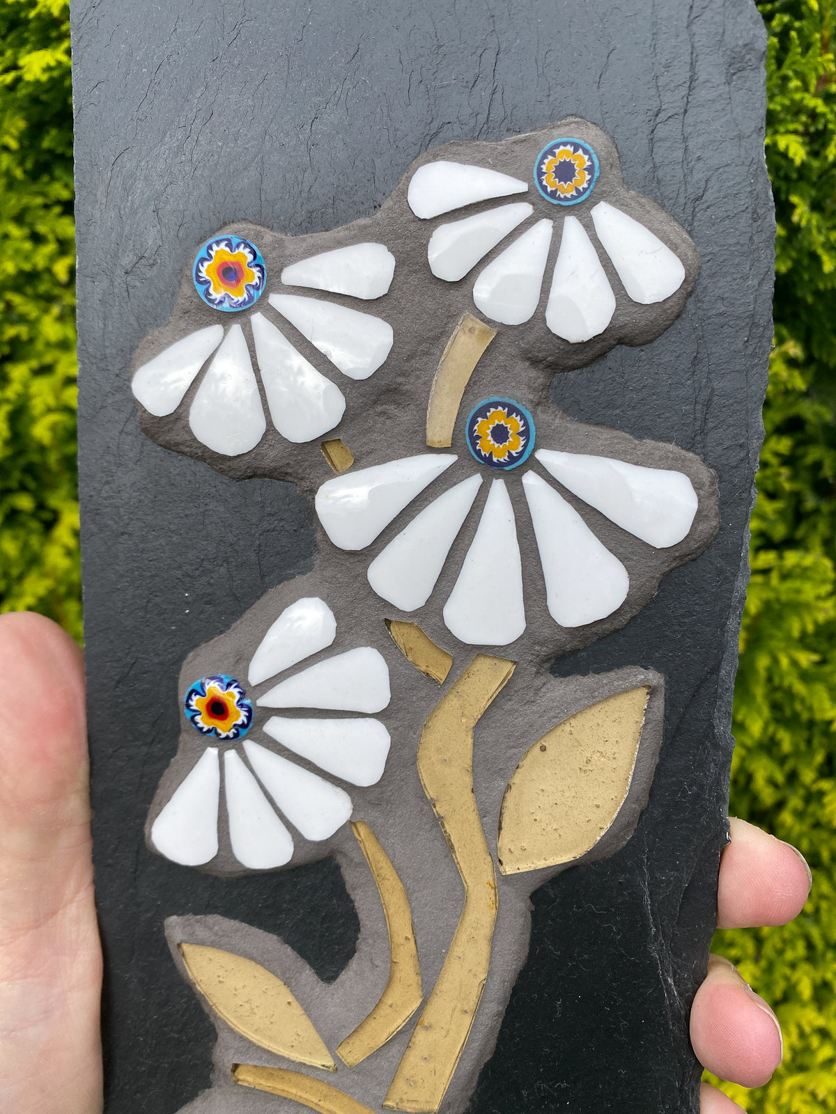 close up of four white daisy mosaics set on dark slate, with tan stems and leaves. Multicoloured centres in blue yellow ans red