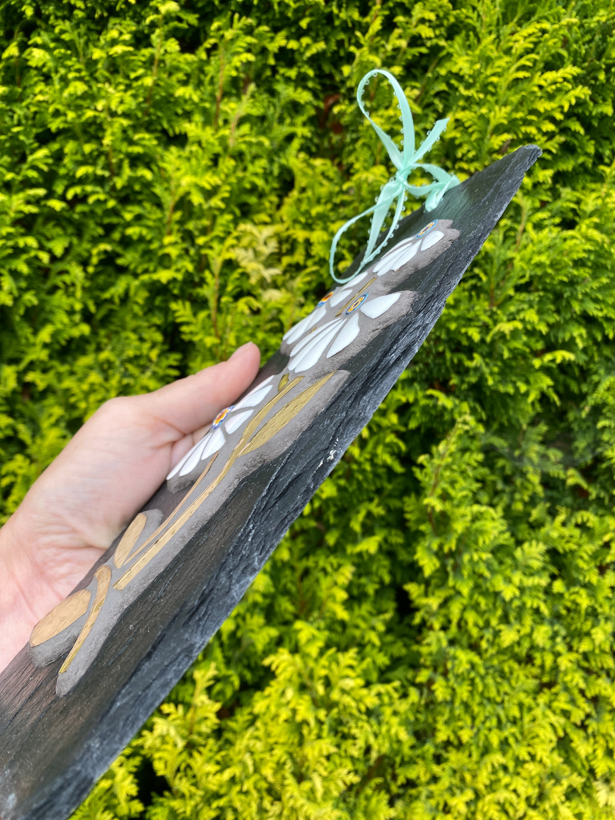 A hand holds a dark slate wall plaque featuring white flowers mosaic, yellow accents, and a mint green hanging loop, set against a background of green foliage.