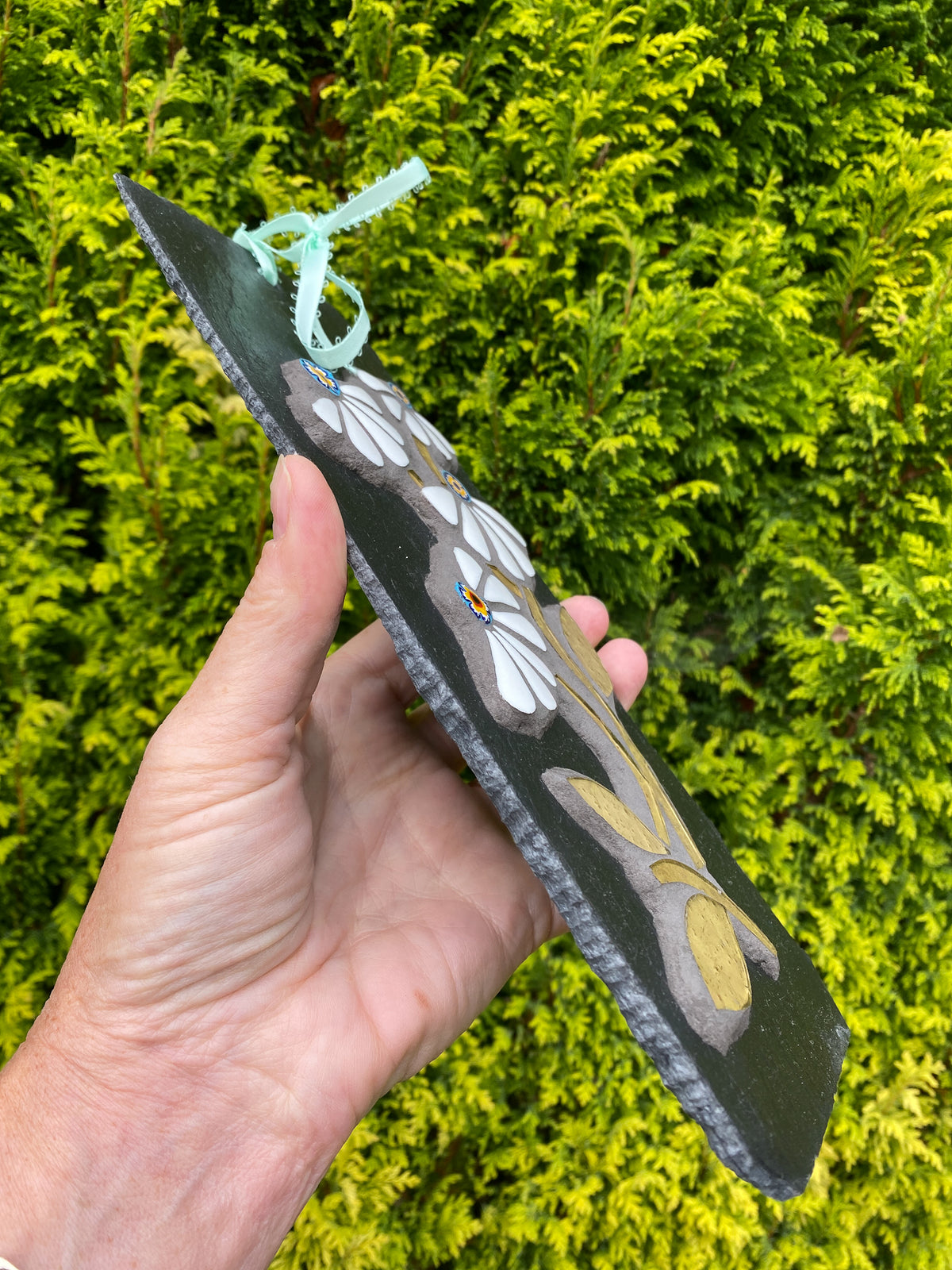 A hand holds a dark slate wall plaque featuring white flowers mosaic, yellow accents, and a mint green hanging loop, set against a background of green foliage.