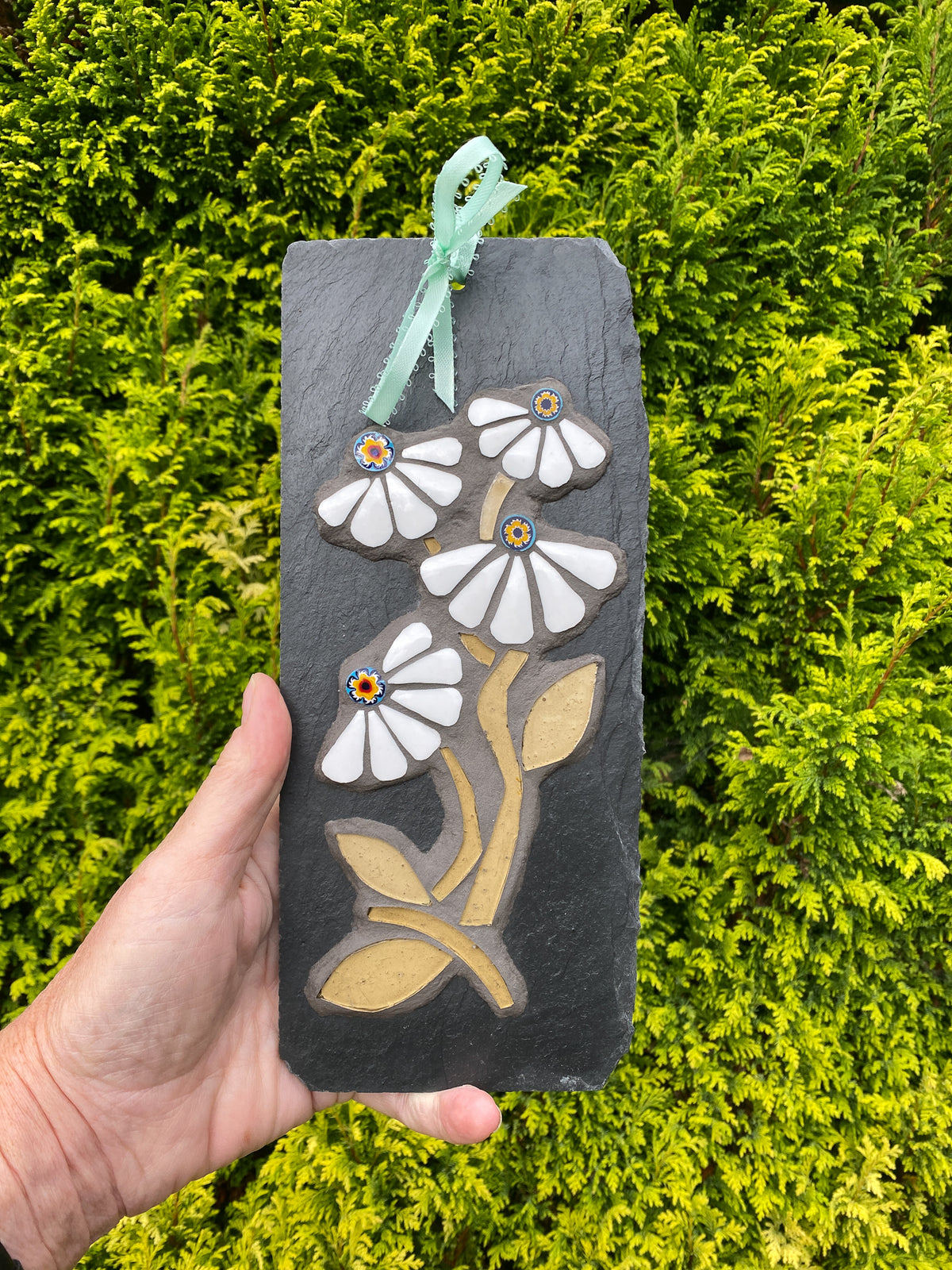 A hand holds a rectangular dark slate plaque featuring a mosaic design of white daisy-like flowers with tan stems and leaves, accented with small colorful centers, and a mint green ribbon loop at the top. The background is a dense green hedge.