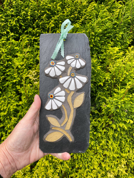 A hand holds a rectangular dark slate plaque featuring a mosaic design of white daisy-like flowers with tan stems and leaves, accented with small colorful centers, and a mint green ribbon loop at the top. The background is a dense green hedge.
