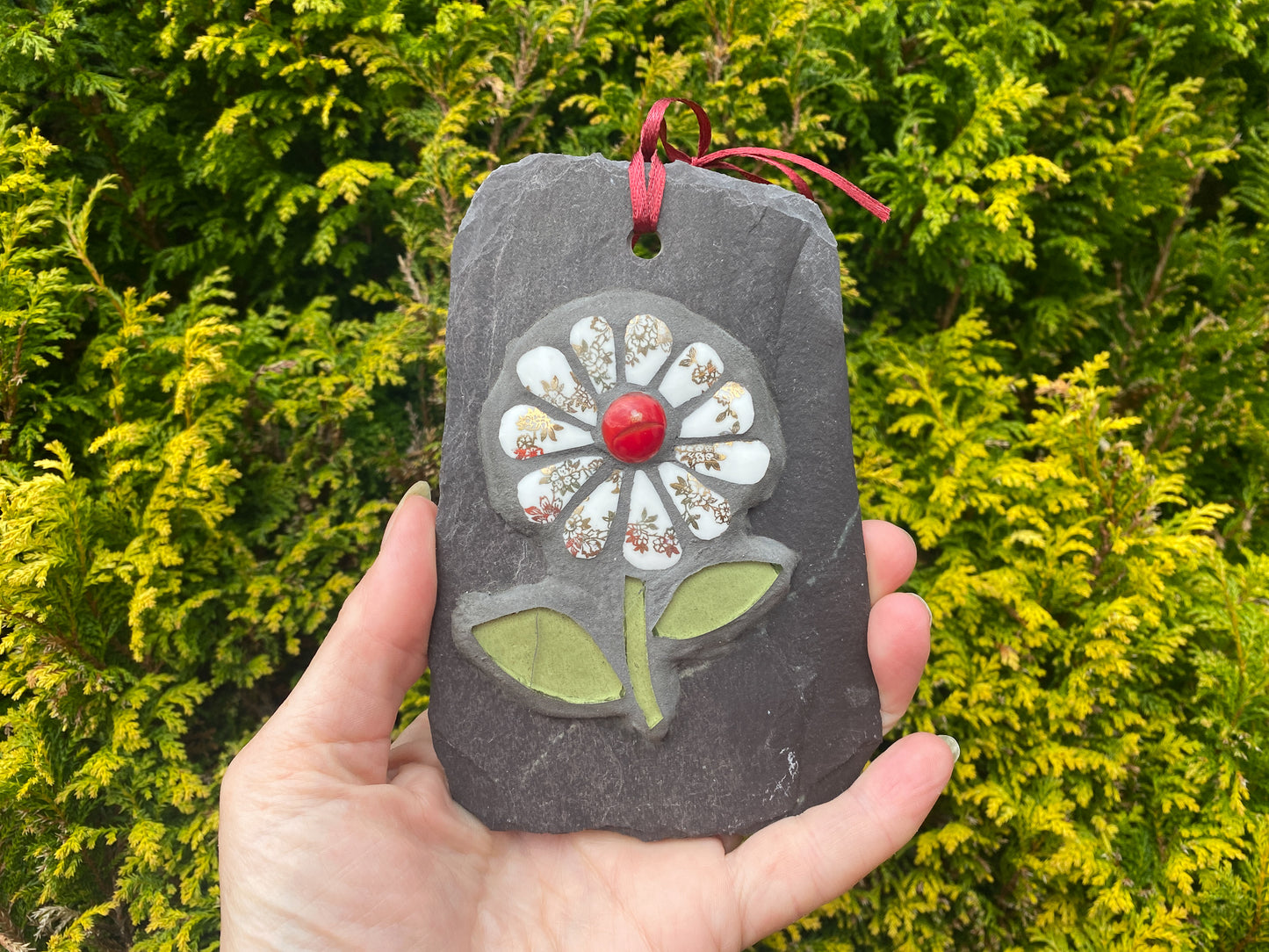 gilt and white petalled daisy mosaic on slate, hand held with an evergreen tree behind