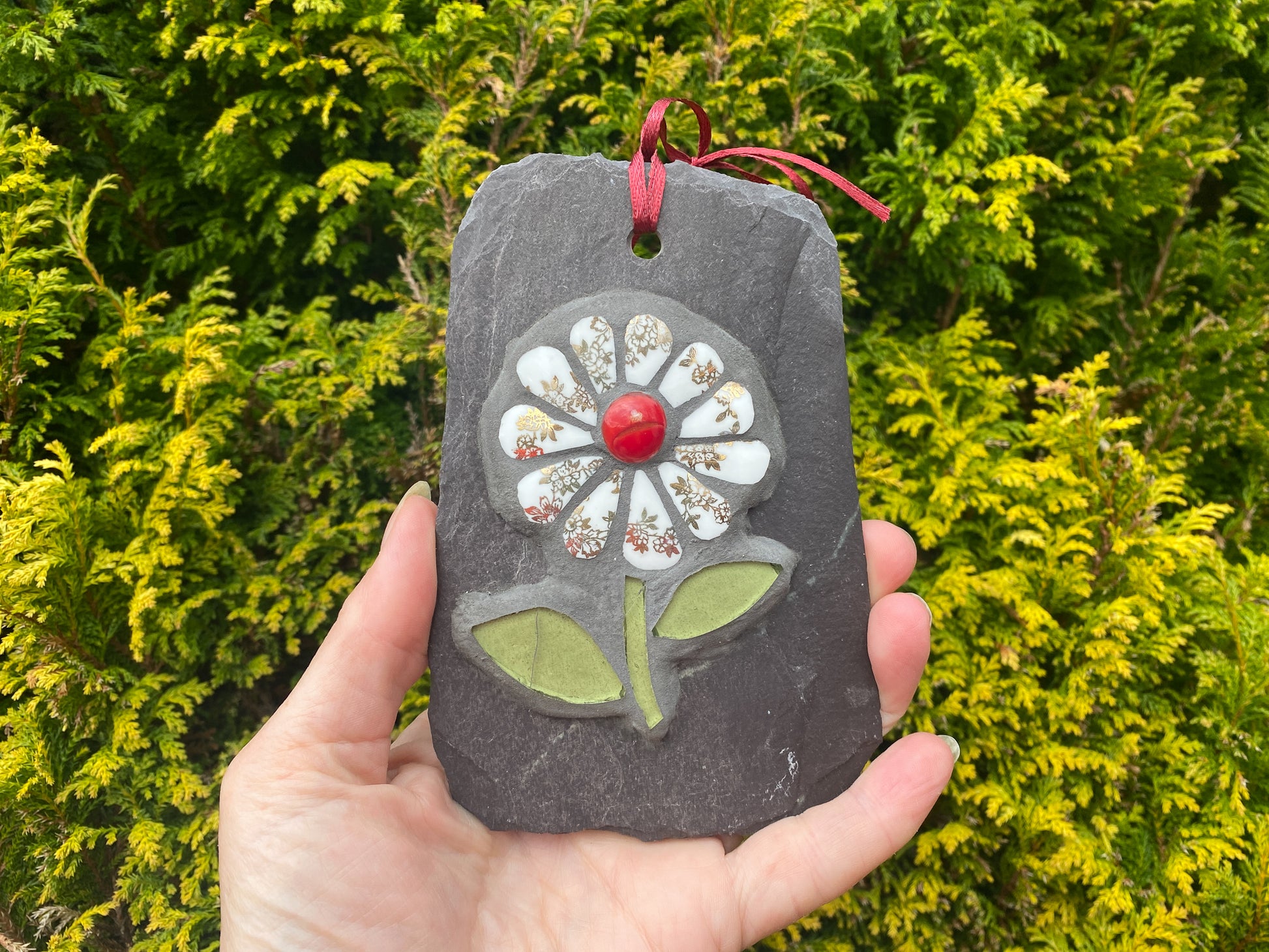 gilt and white petalled daisy mosaic on slate, hand held with an evergreen tree behind